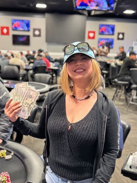 Smiling person in a cap holds a fan of cash at a poker table in a busy casino room