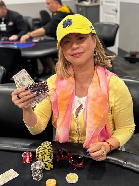 Woman at a poker table holding cash and chips, wearing a yellow cap and pink scarf.