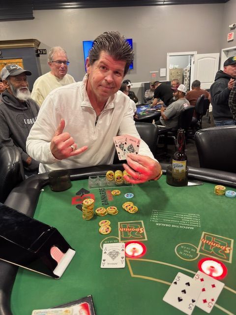 Man in white shirt at a casino table, gesturing toward chips and cards during a card game.
