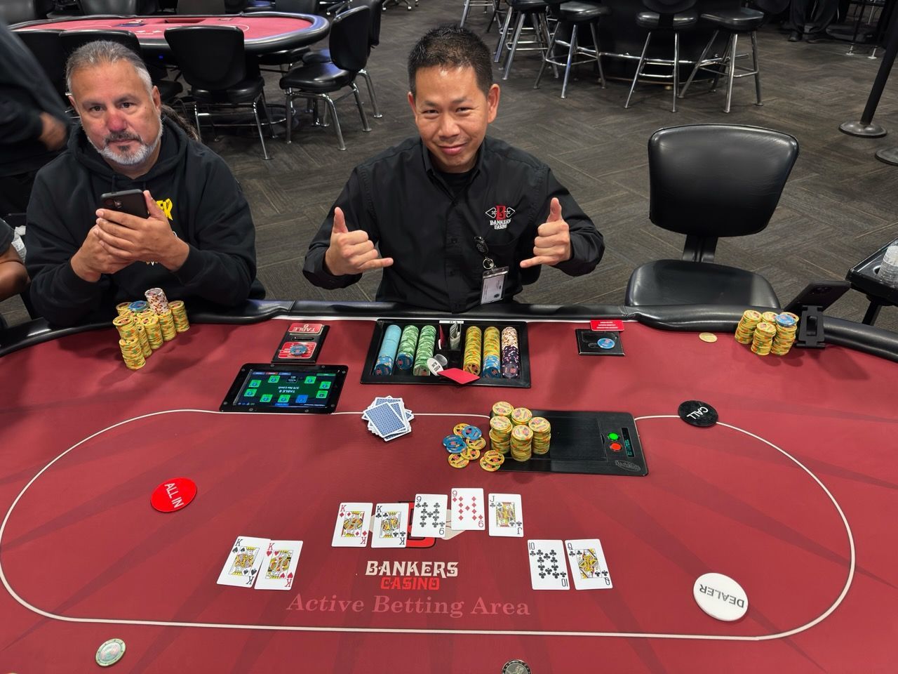 Two men sitting at a casino table, smiling with cards, chips, and poker game equipment in front of them