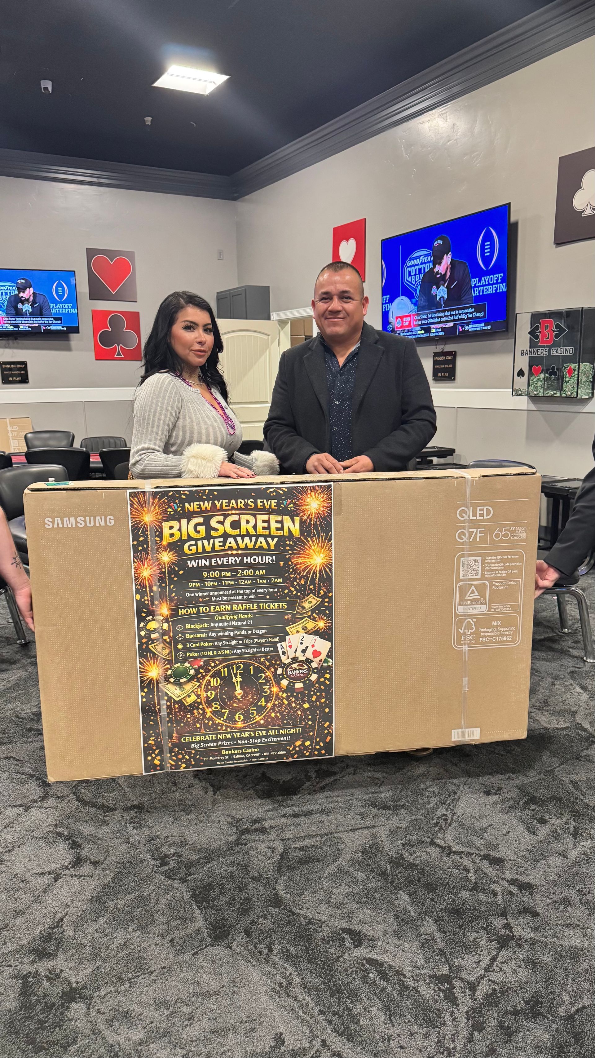 Two people standing behind a large cardboard box in a showroom with multiple TVs on the wall.
