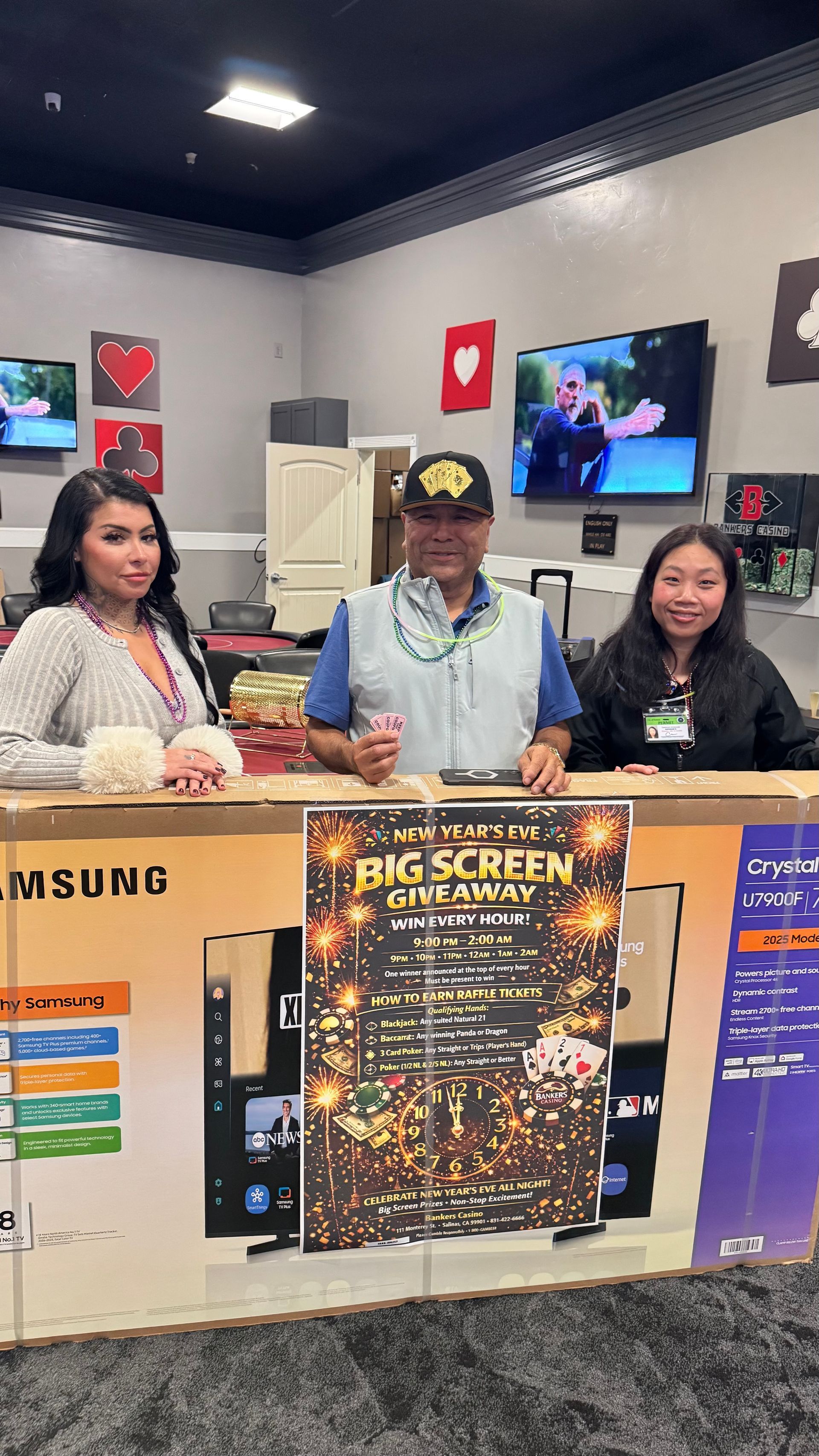 Three people posing behind a Samsung display at a gaming store, with a TV and promotional posters behind them.