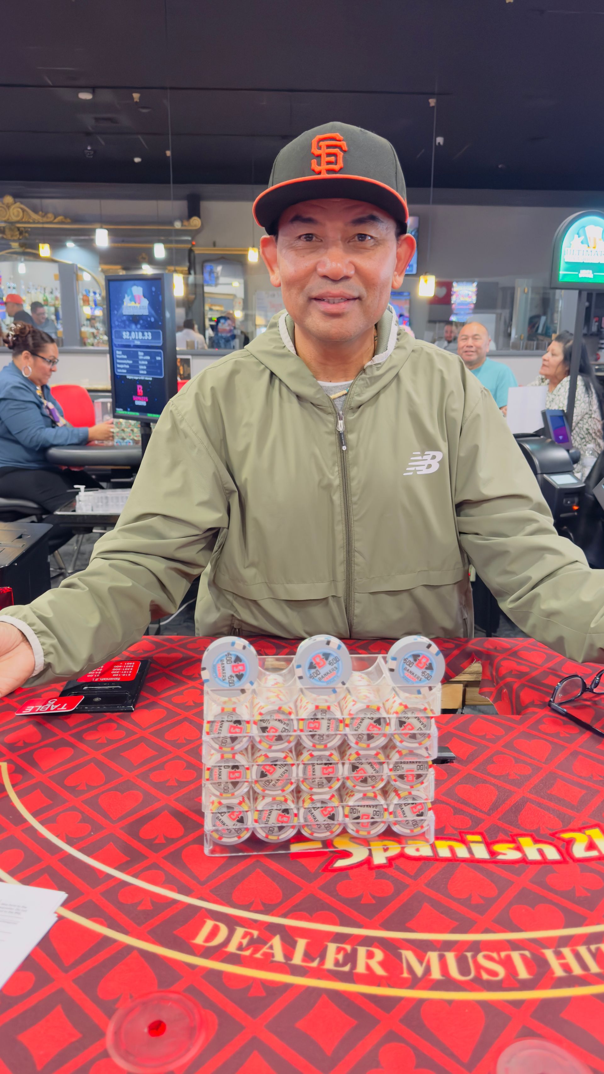 Person standing at a casino table with stacks of chips, wearing a green jacket and black cap.