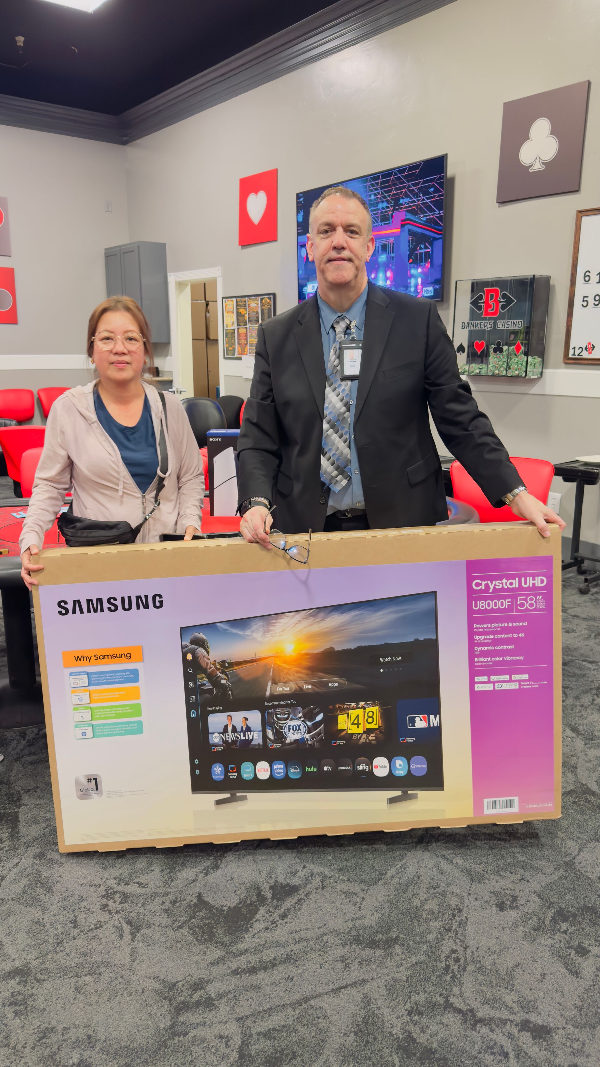Two people standing behind a boxed Samsung TV in a store, with gaming chairs and wall decor in the background
