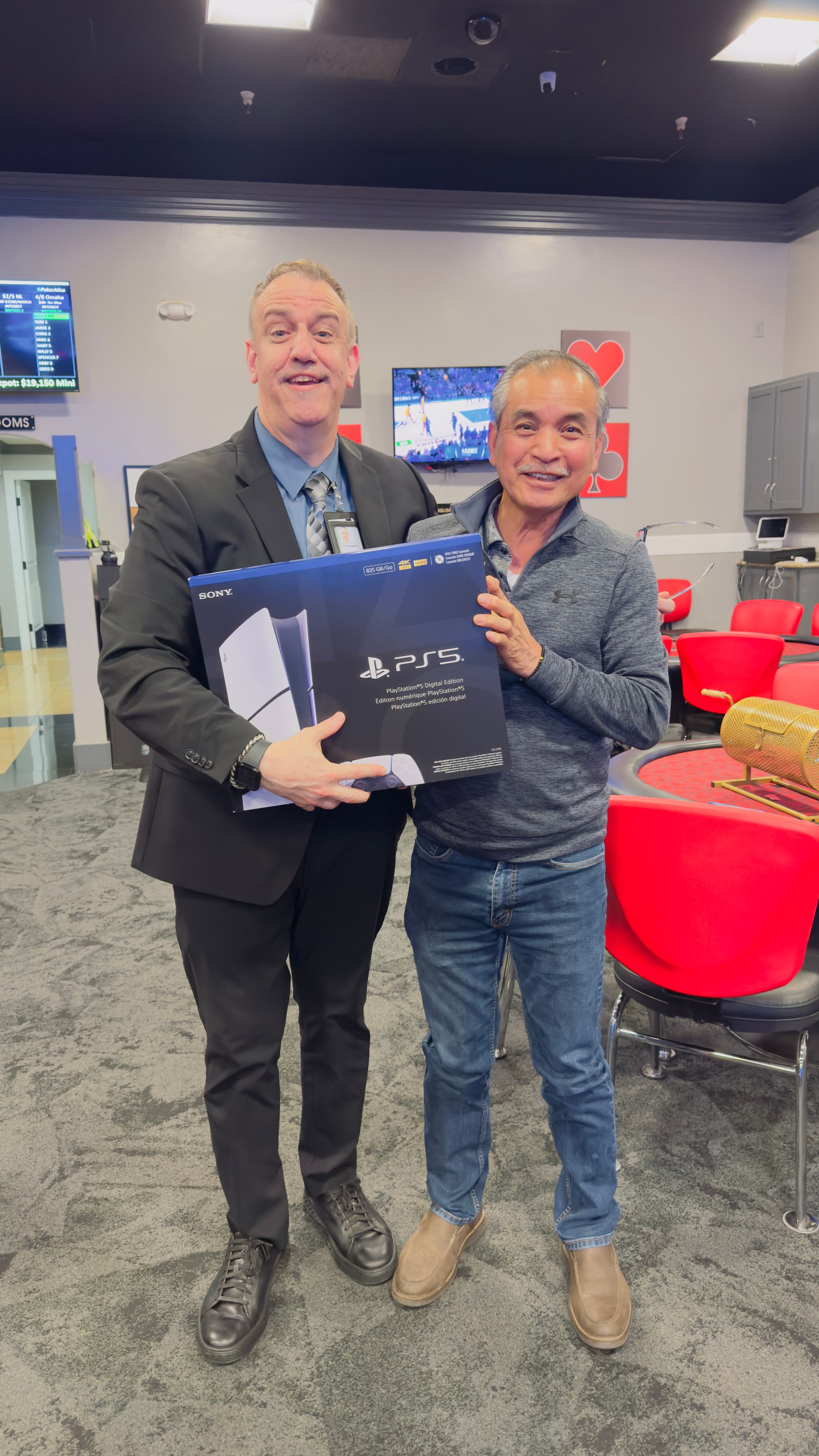 Two men posing indoors, smiling and holding a boxed item in a room with red chairs.