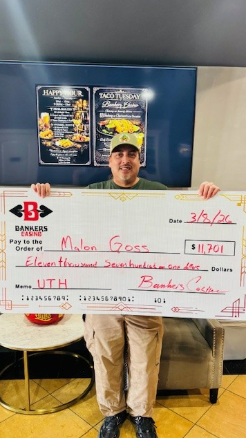 Man holding oversized check in front of sponsor banners at an event, smiling and wearing a cap.