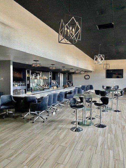 Modern bar lounge with black stools, wood flooring, and pendant lights under a dark ceiling.