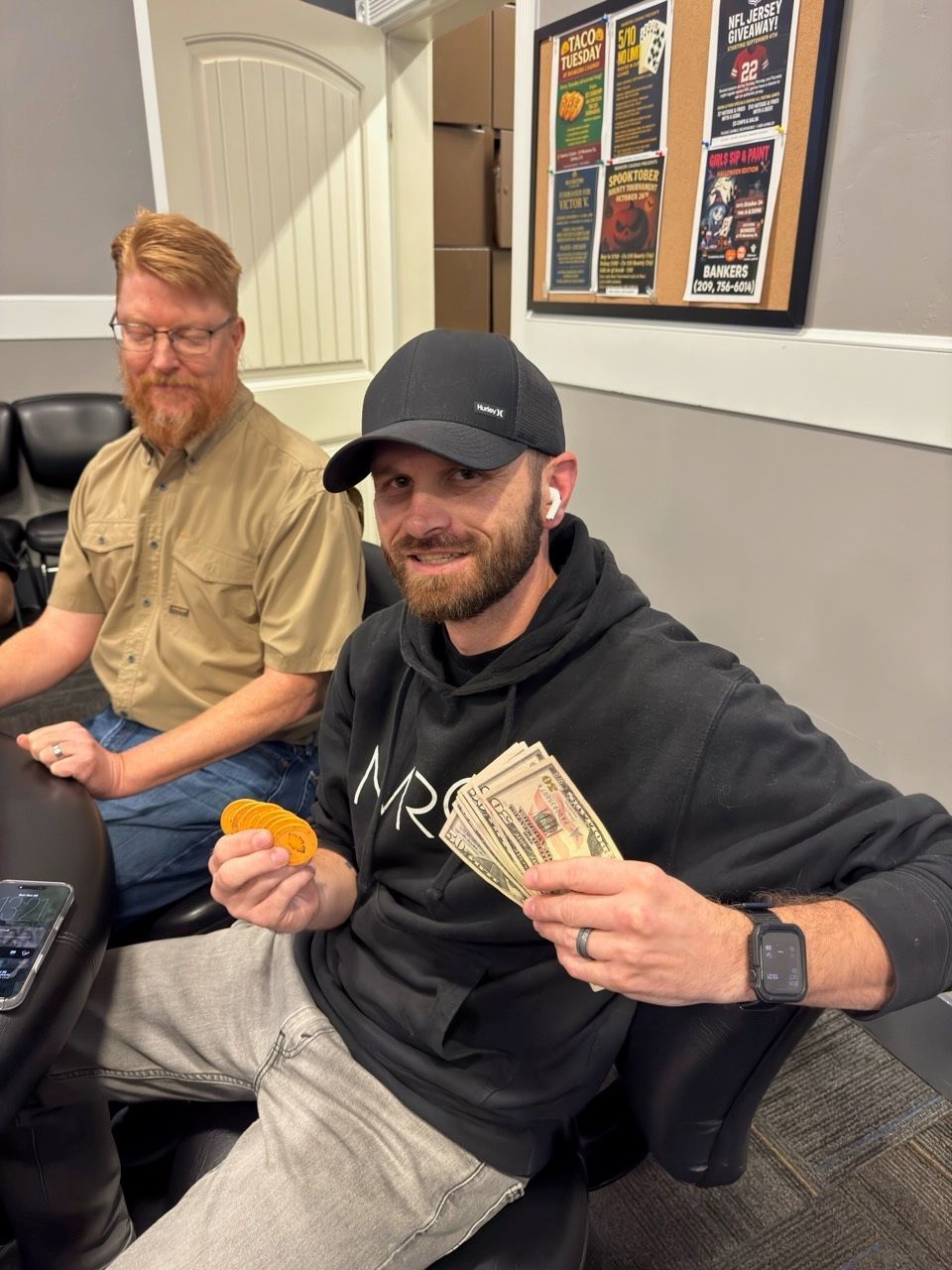 Two men on a couch, one holding money and an orange snack, in a casual indoor room.