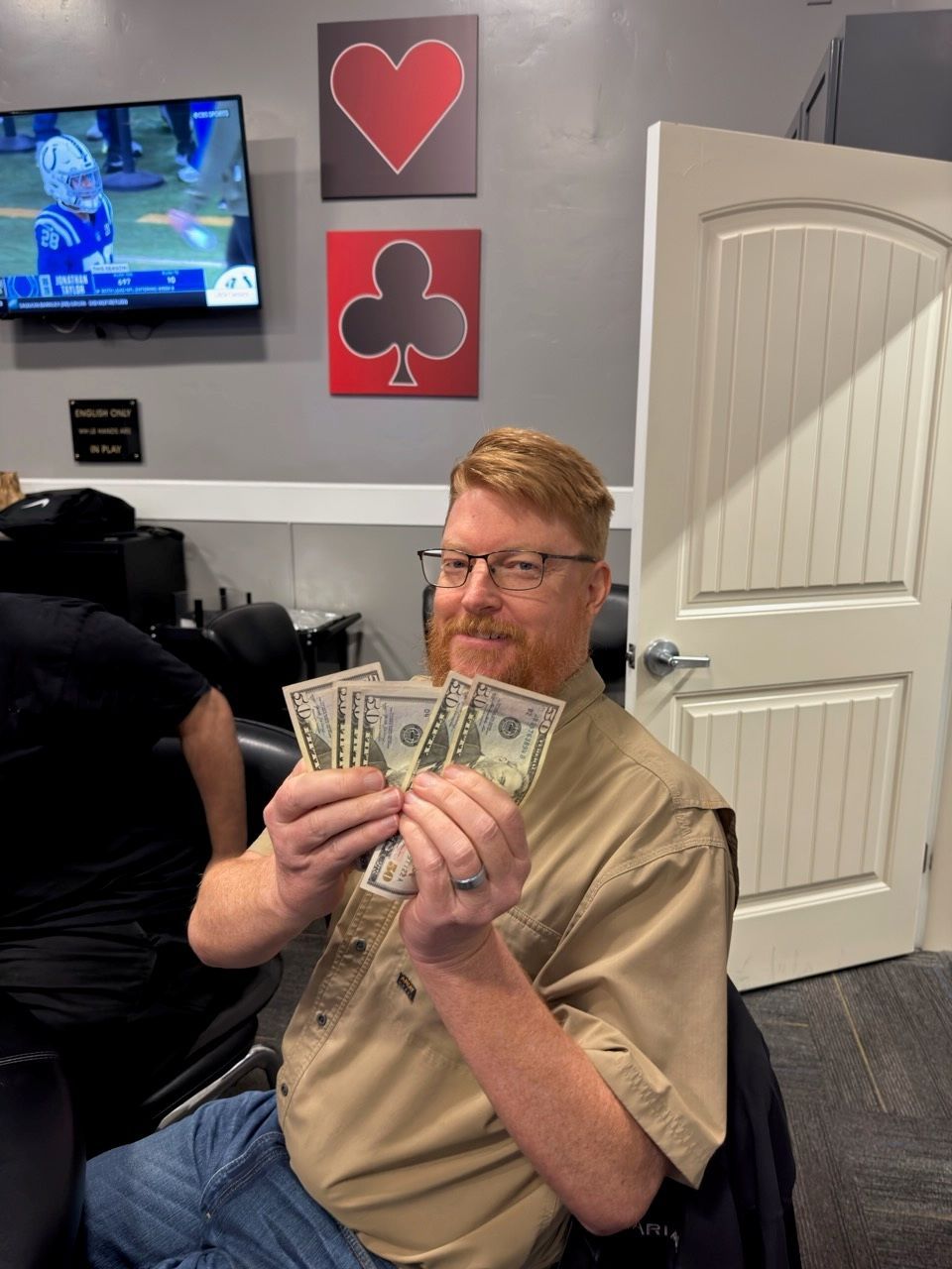 Man smiling while holding a fan of cash in a room with card-suit wall art and a TV.