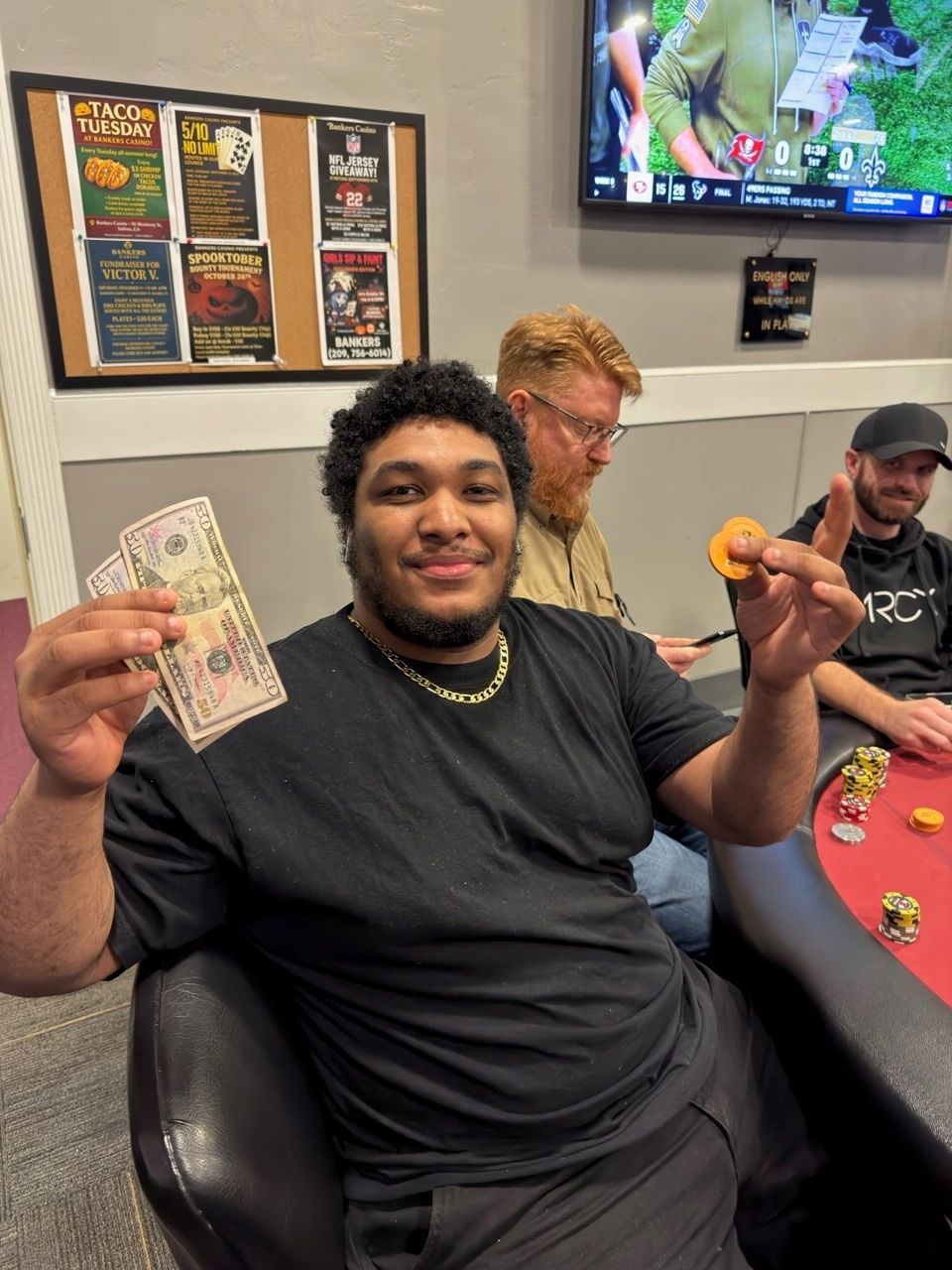 Man holding cash at a casino table, smiling with two people seated behind him.