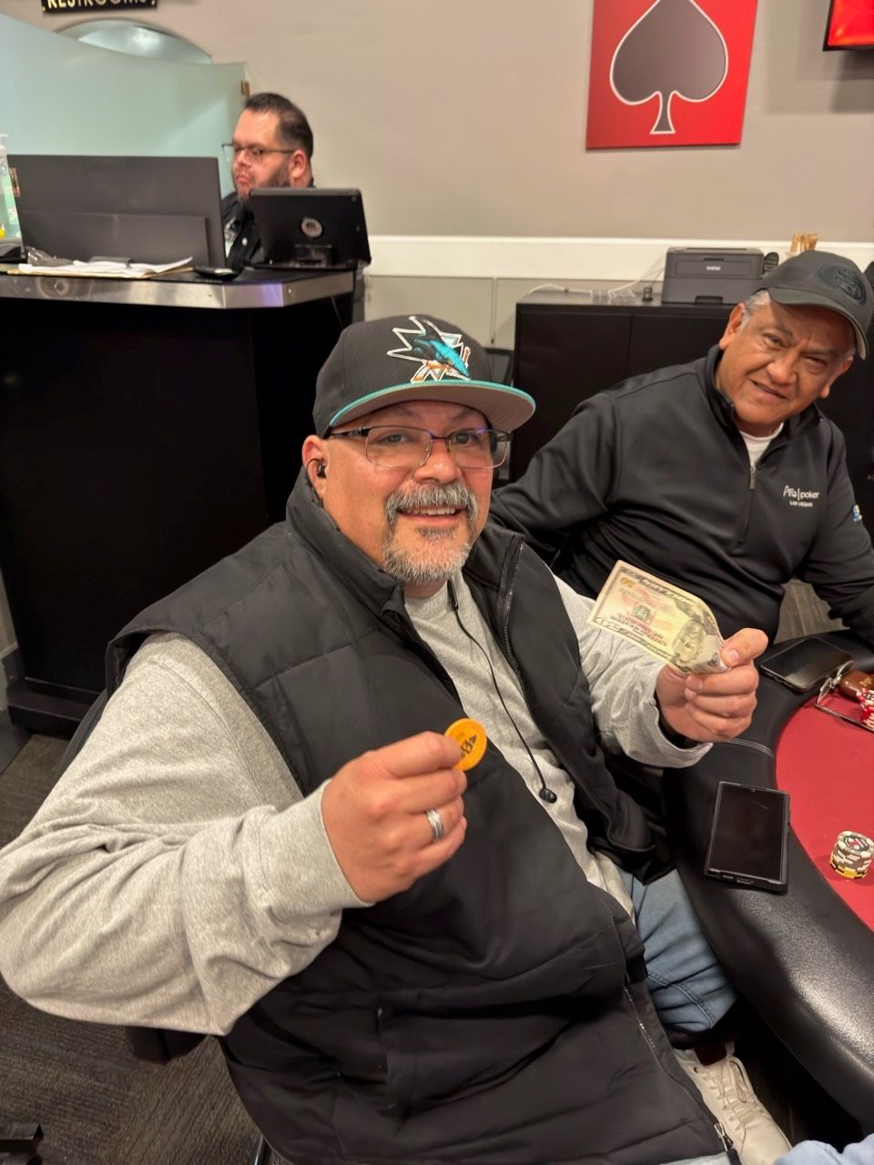 Two men smiling at a restaurant table, one holding a small snack and the other holding cash.