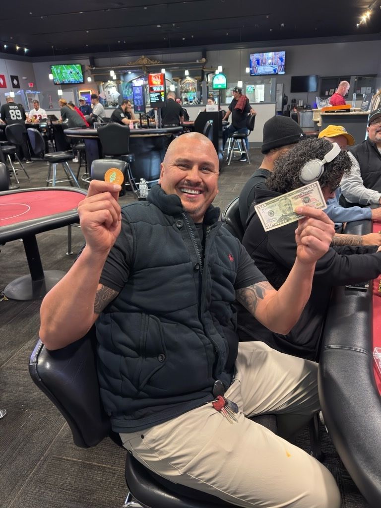 Smiling man in a black vest sits in a casino, holding up cash and a small orange chip.