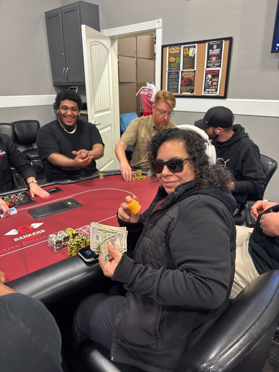 People seated around a red poker table, smiling and holding cards and drinks in a game room.