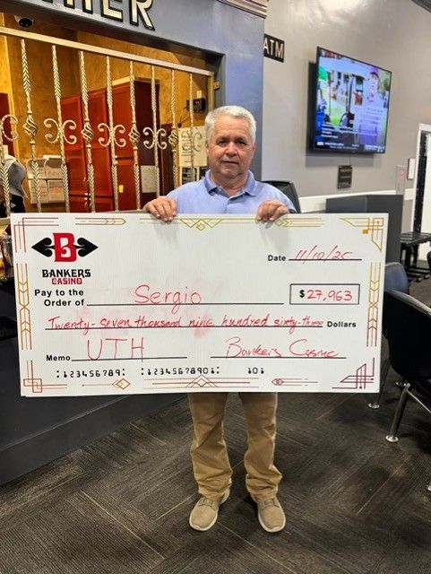 Man holding a large ceremonial check in a lobby, with a TV and signage in the background.