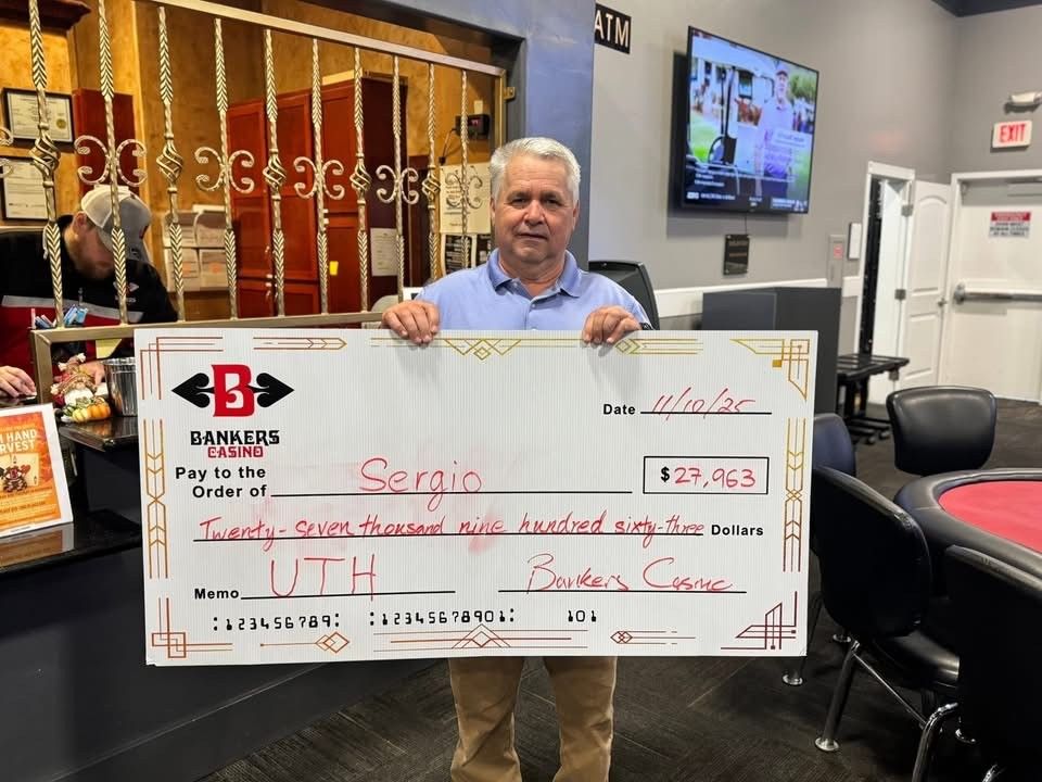 Man holding oversized check in a B. Riley office, with TV and chairs in the background.