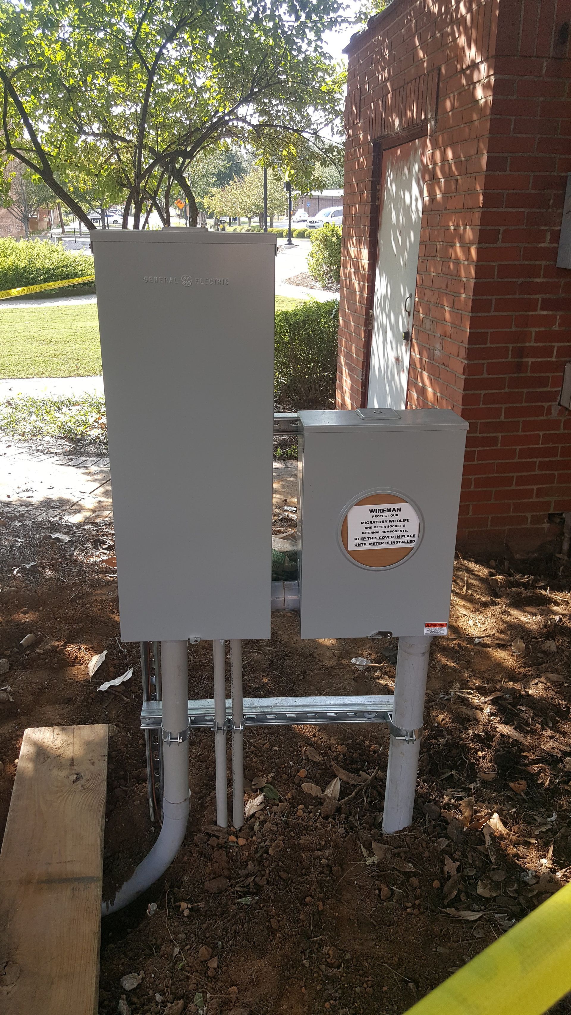 Gray electrical boxes and conduit on a construction site with a red brick wall and yellow caution tape.