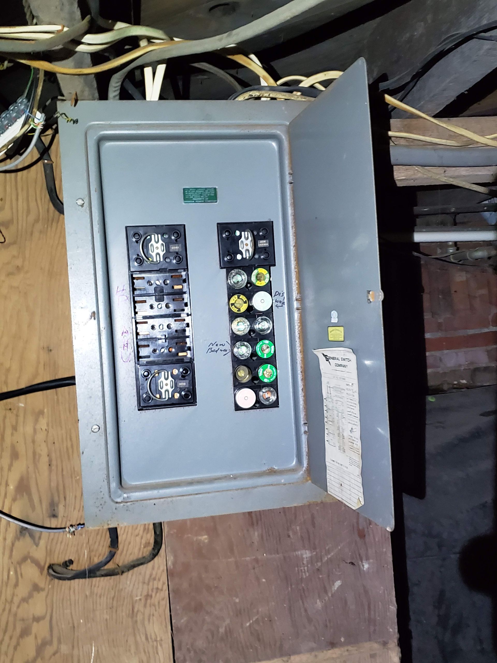 Electrical panel with breakers and fuses, open, mounted on wood in a basement.