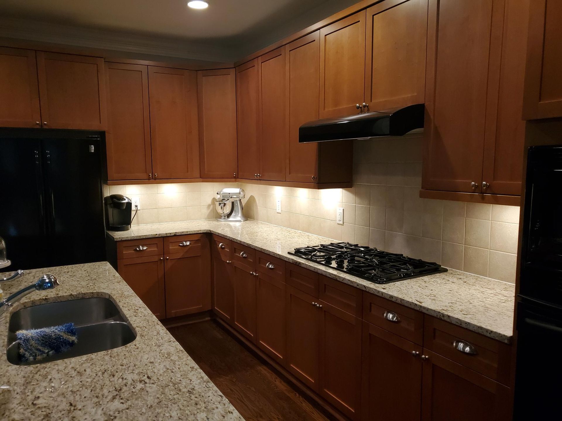 Warm kitchen with wood cabinets, granite counters, black appliances, and under-cabinet lighting.