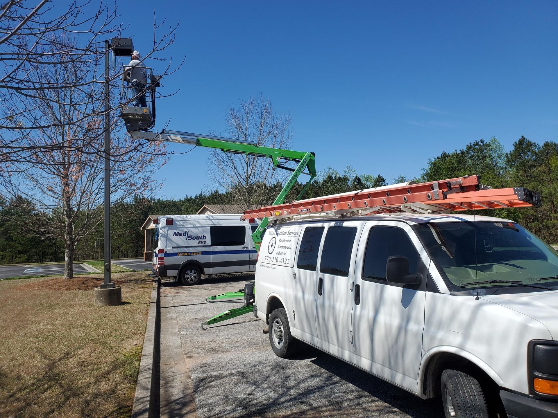 Utility worker in bucket lift trimming tree branches beside parked white vans on a sunny street