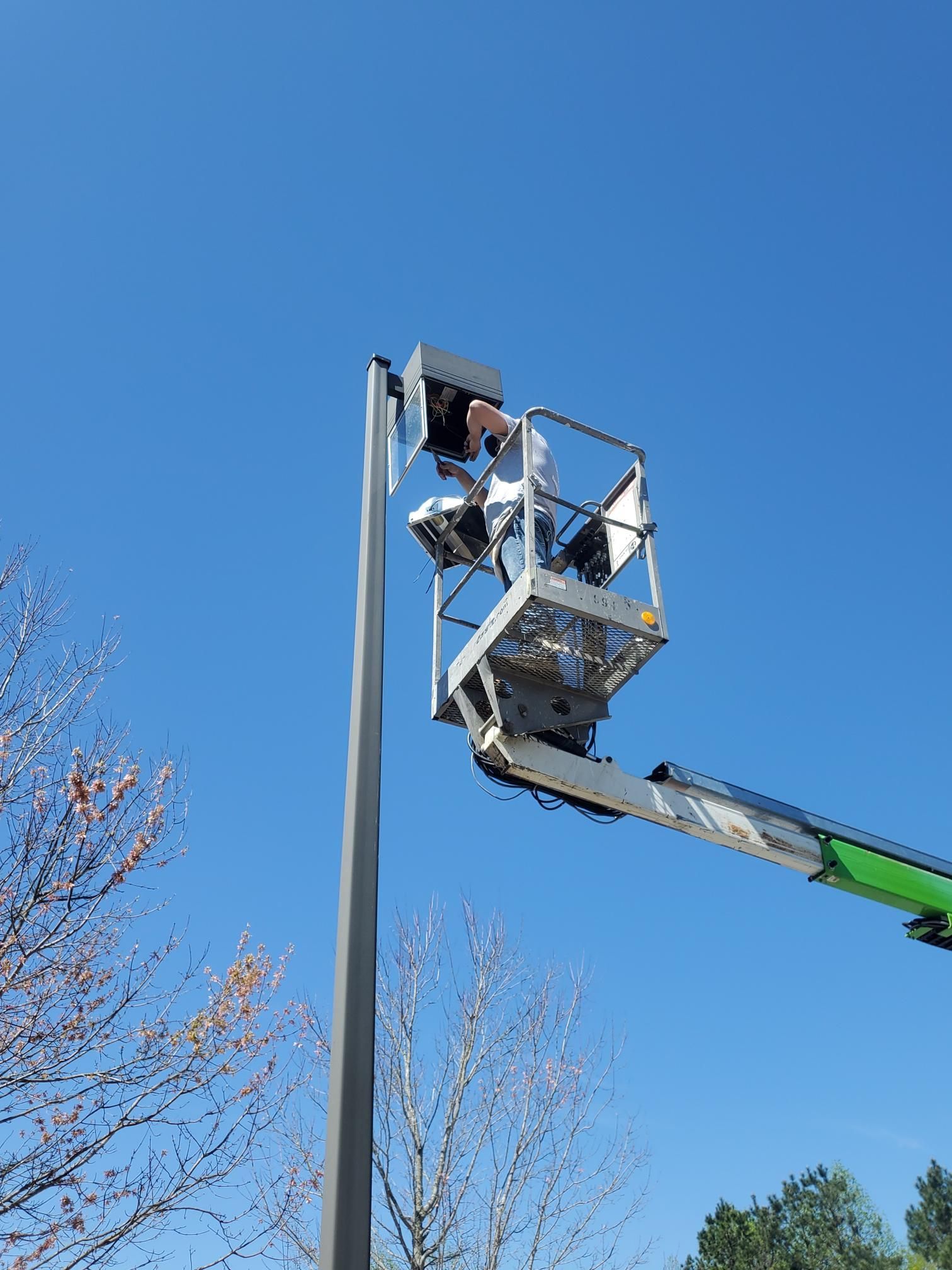 Two workers in a lift repairing a tall streetlight against a clear blue sky