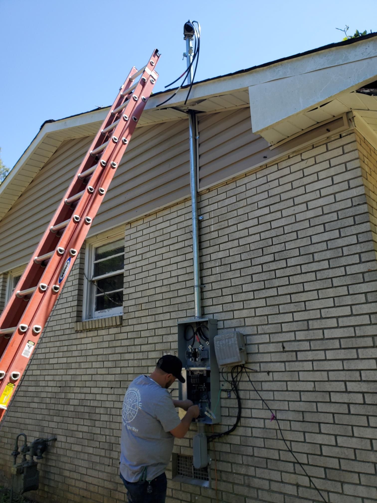 Man working on an exterior house electrical panel beside a red ladder under a roofline