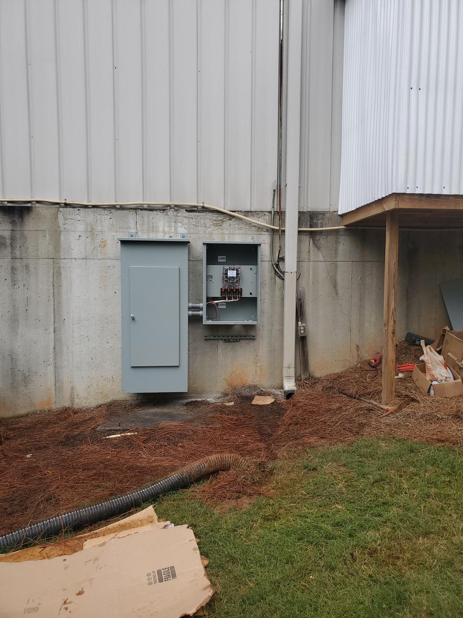 Utility boxes on a concrete wall beside a building, with grass and a hose in the foreground.