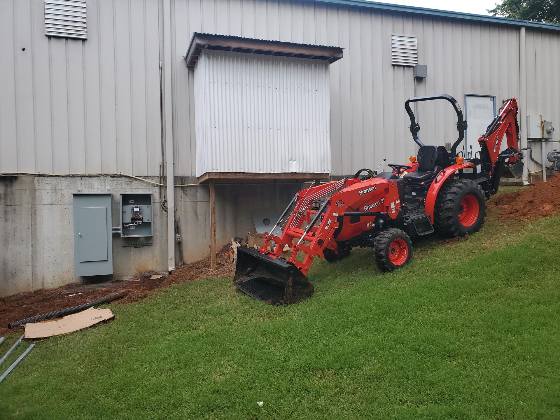 Red tractor with front loader and backhoe parked on a grassy slope beside a gray building