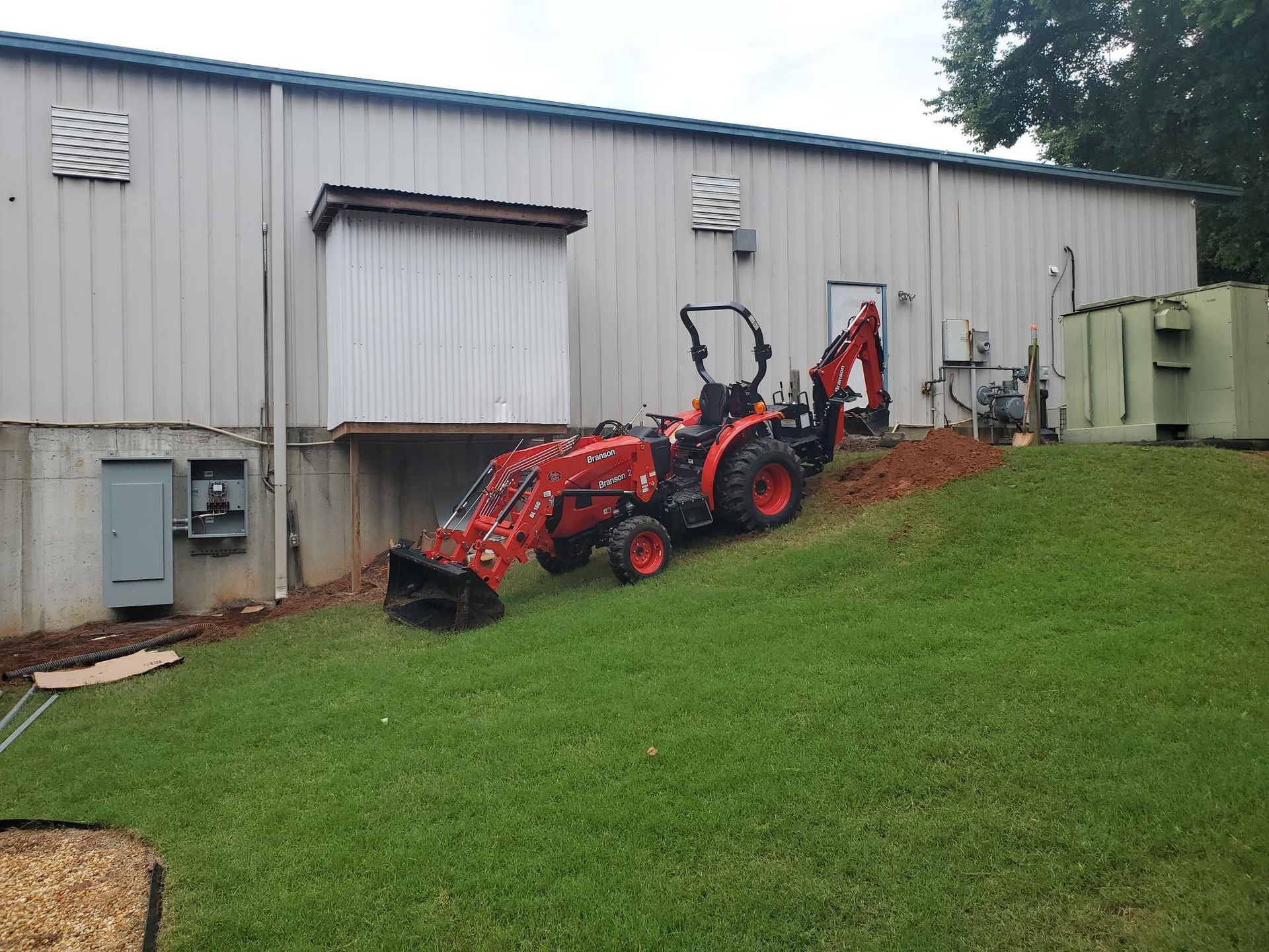 Red tractor and backhoe parked beside a gray building on a grassy slope