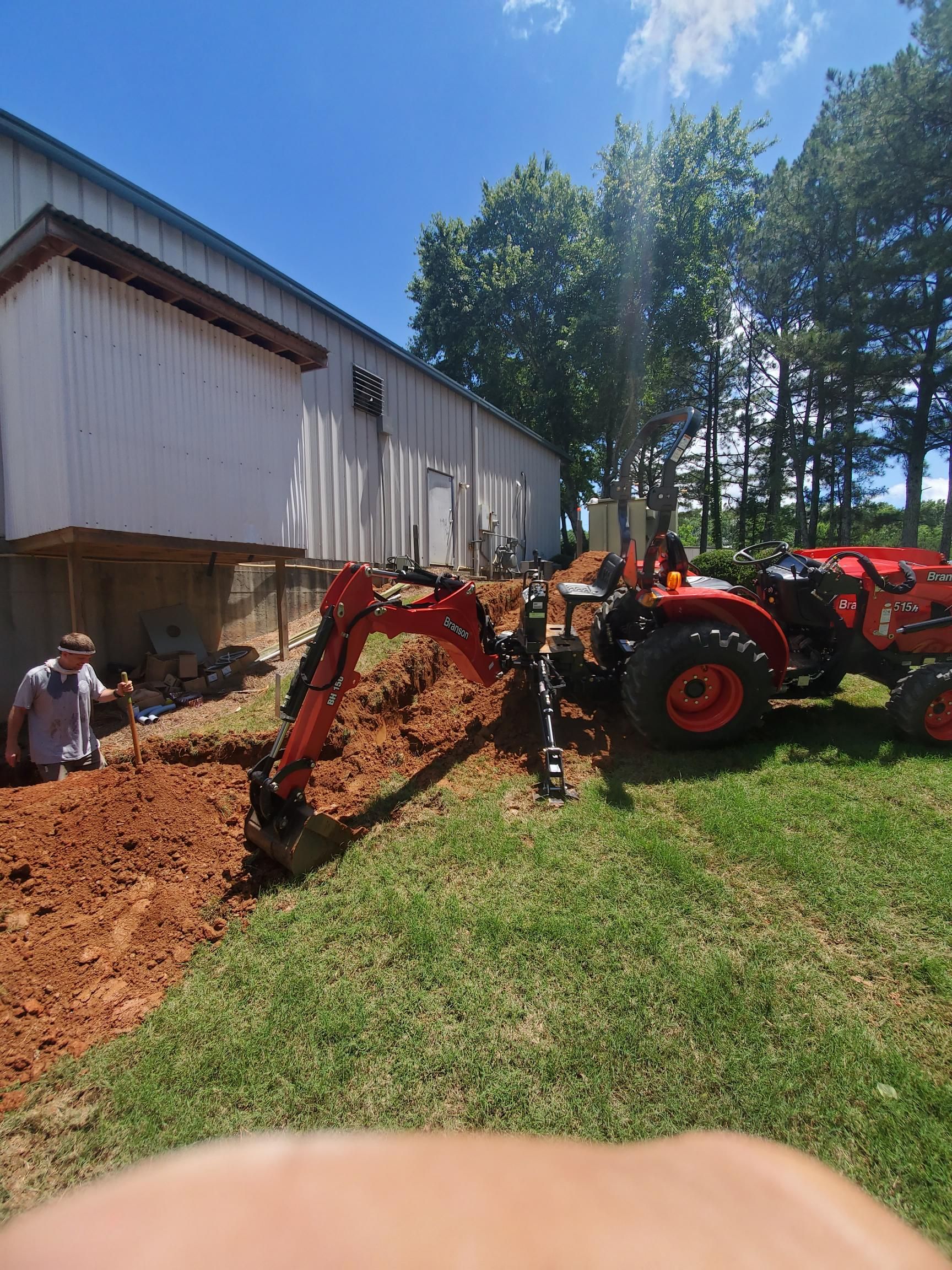 Red tractor on dirt trench beside a metal building, with a worker nearby and trees in the background