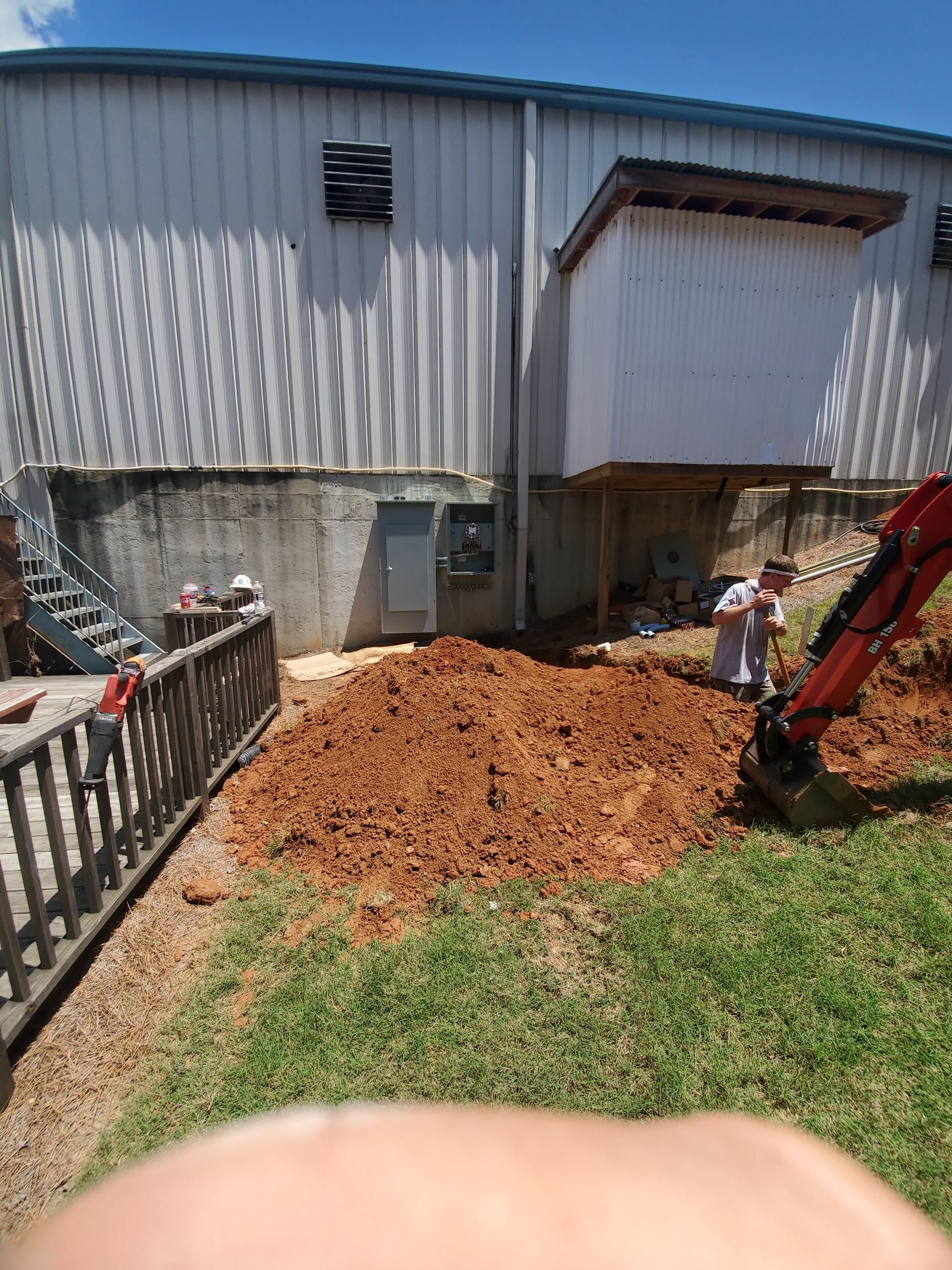 Backyard soil pile beside a building, with a small excavator and a person near a fence.