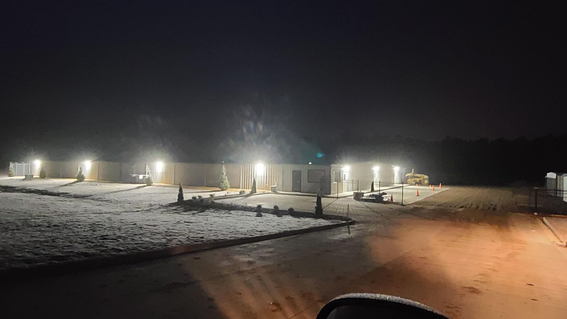 Snowy industrial lot at night, lit by bright lamps under a dark sky.