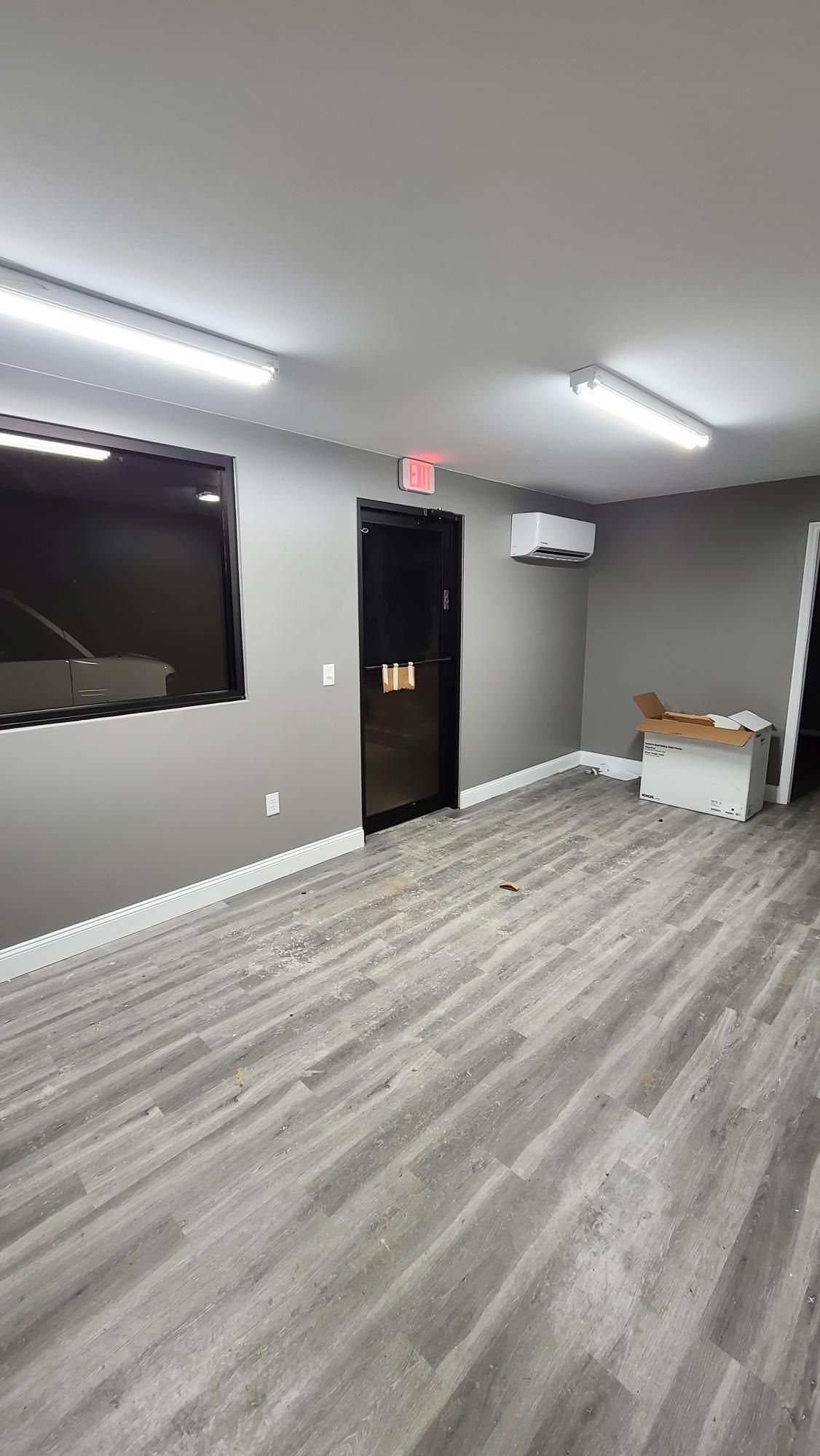 Empty office room with gray wood floors, black door, wall-mounted TV, and fluorescent ceiling lights