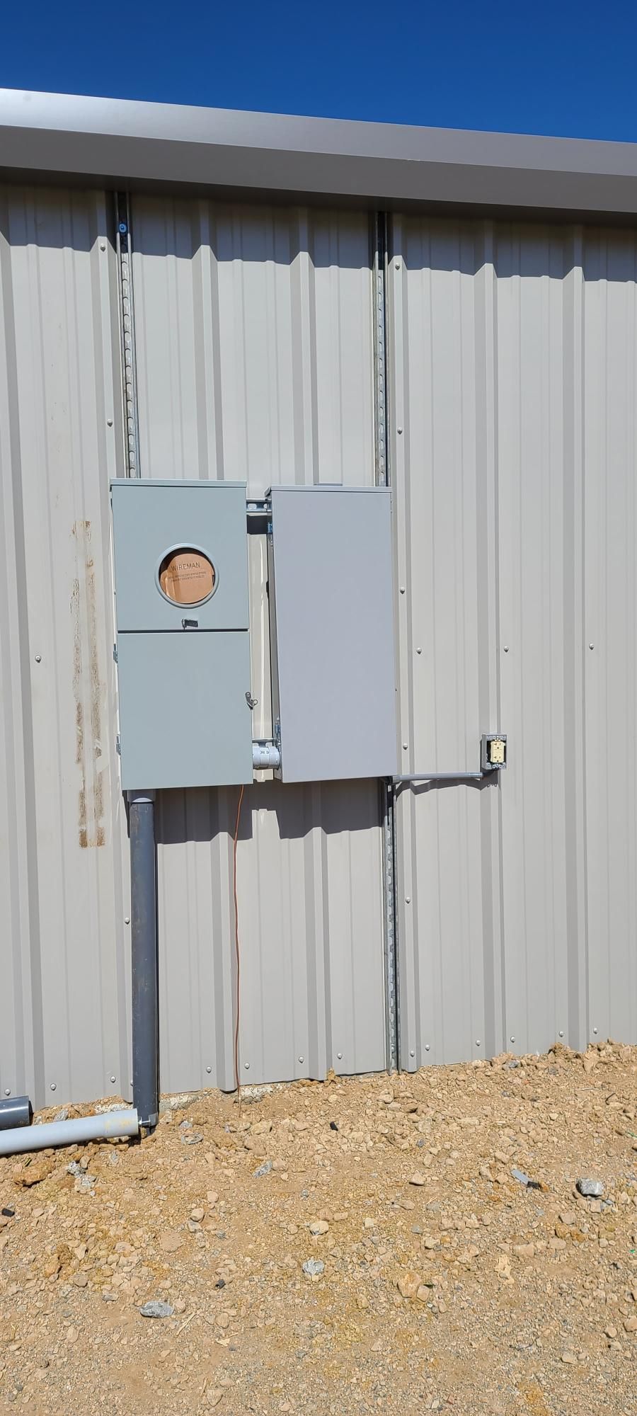 Gray utility boxes mounted on a corrugated metal building wall over gravel ground