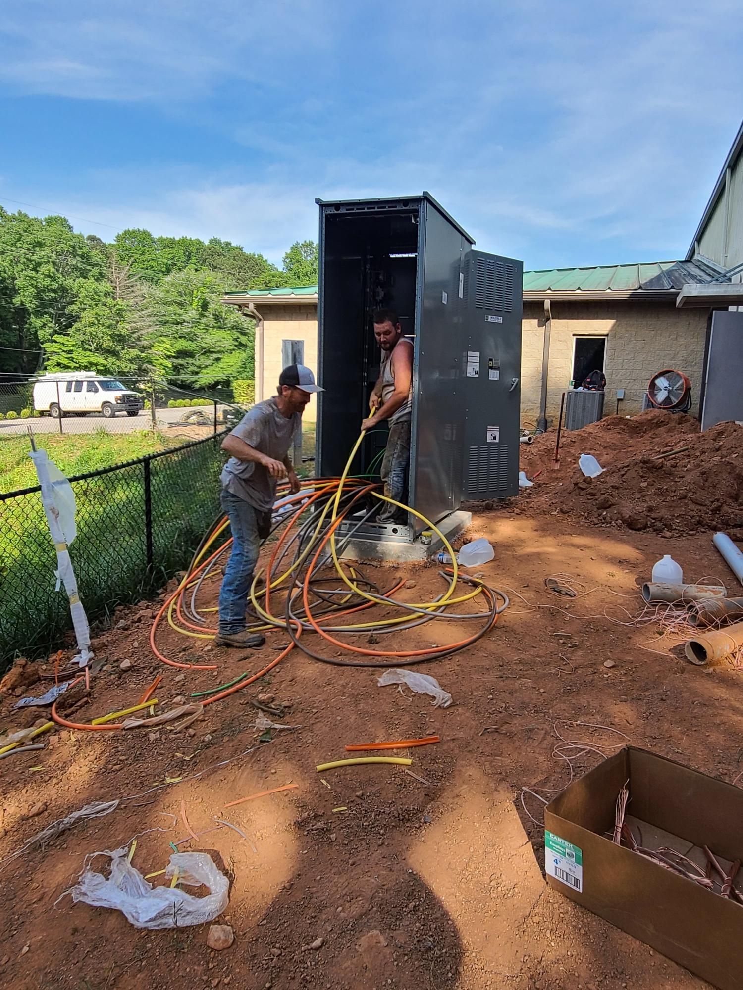 Two workers on a muddy construction site handling cables near a large metal equipment box.