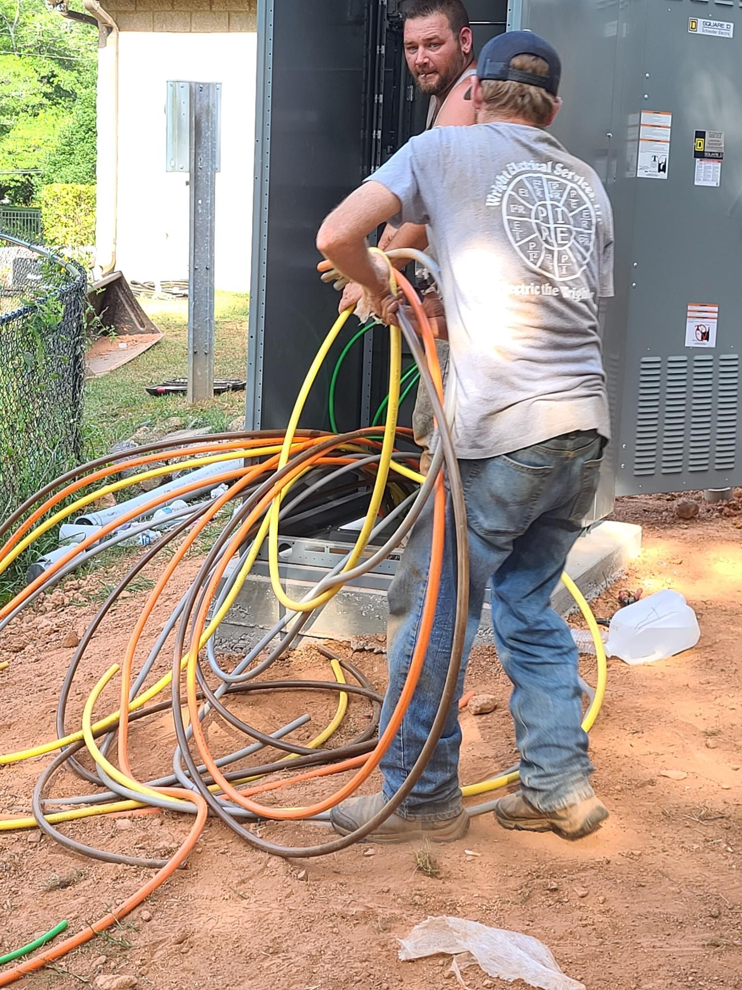 Two workers handling tangled orange, yellow, and blue cables beside a gray utility box outdoors