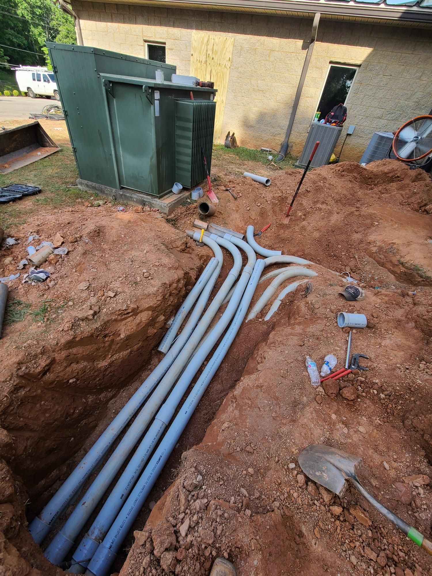 Trenches with gray conduits laid across red dirt beside green utility boxes near a building.