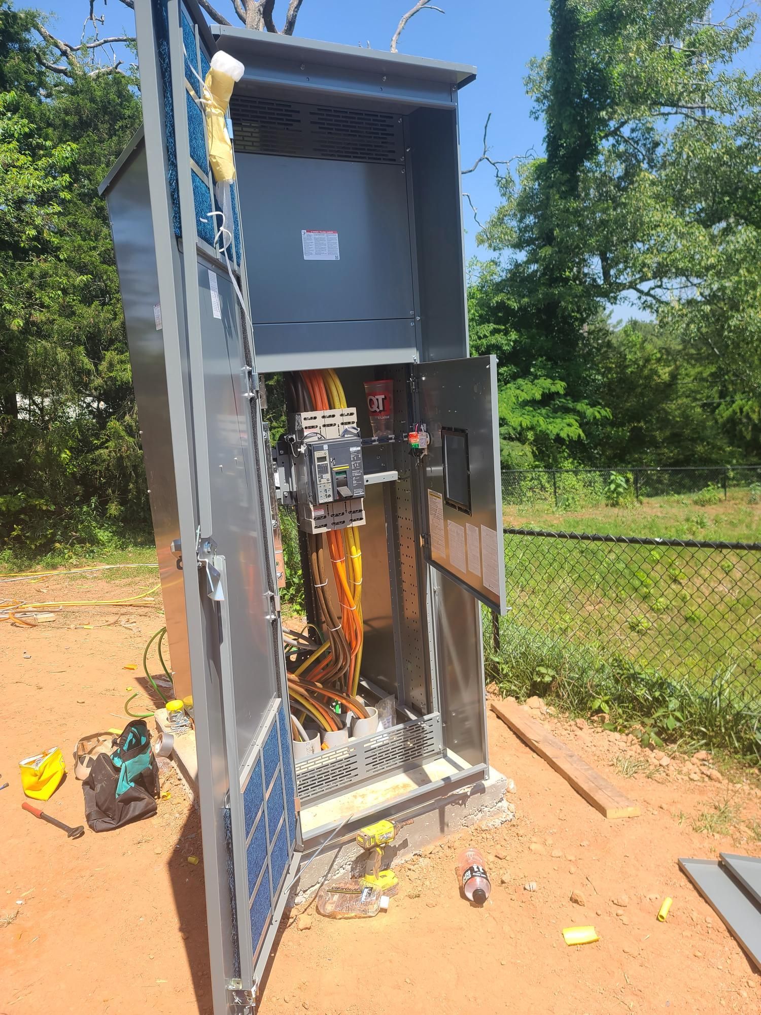 Open electrical cabinet with exposed wiring outdoors by a dirt path and chain-link fence.