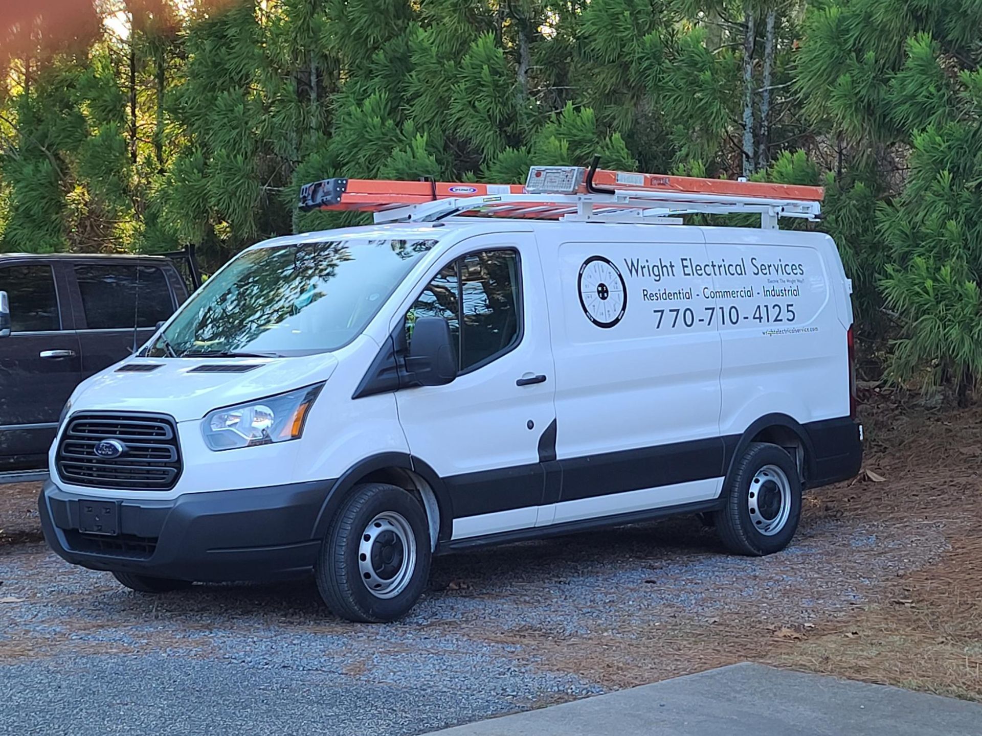 White service van parked on a gravel lot with orange ladders on top, beside a wooded area