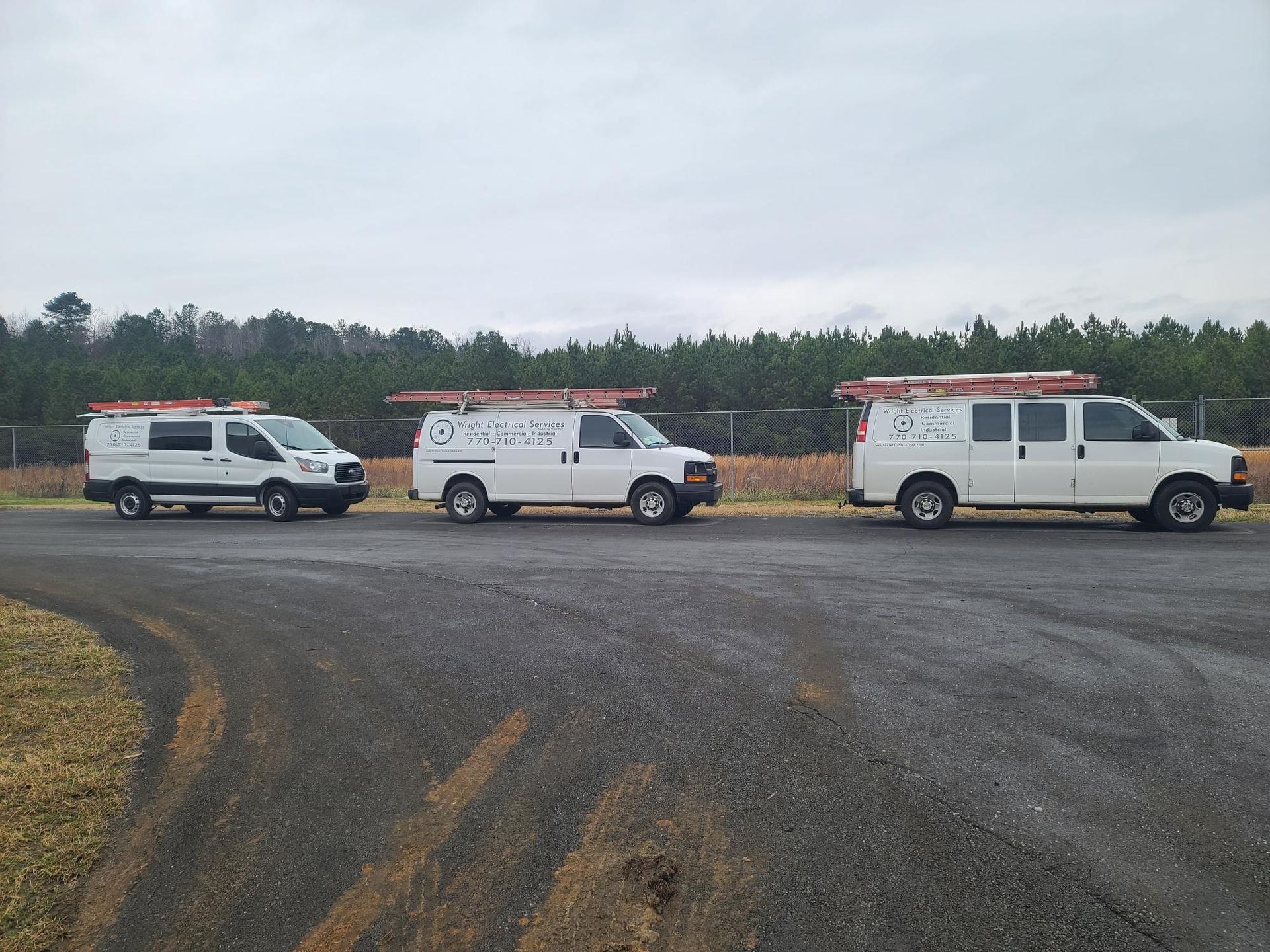 Three white utility vans parked on a gravel lot under an overcast sky.