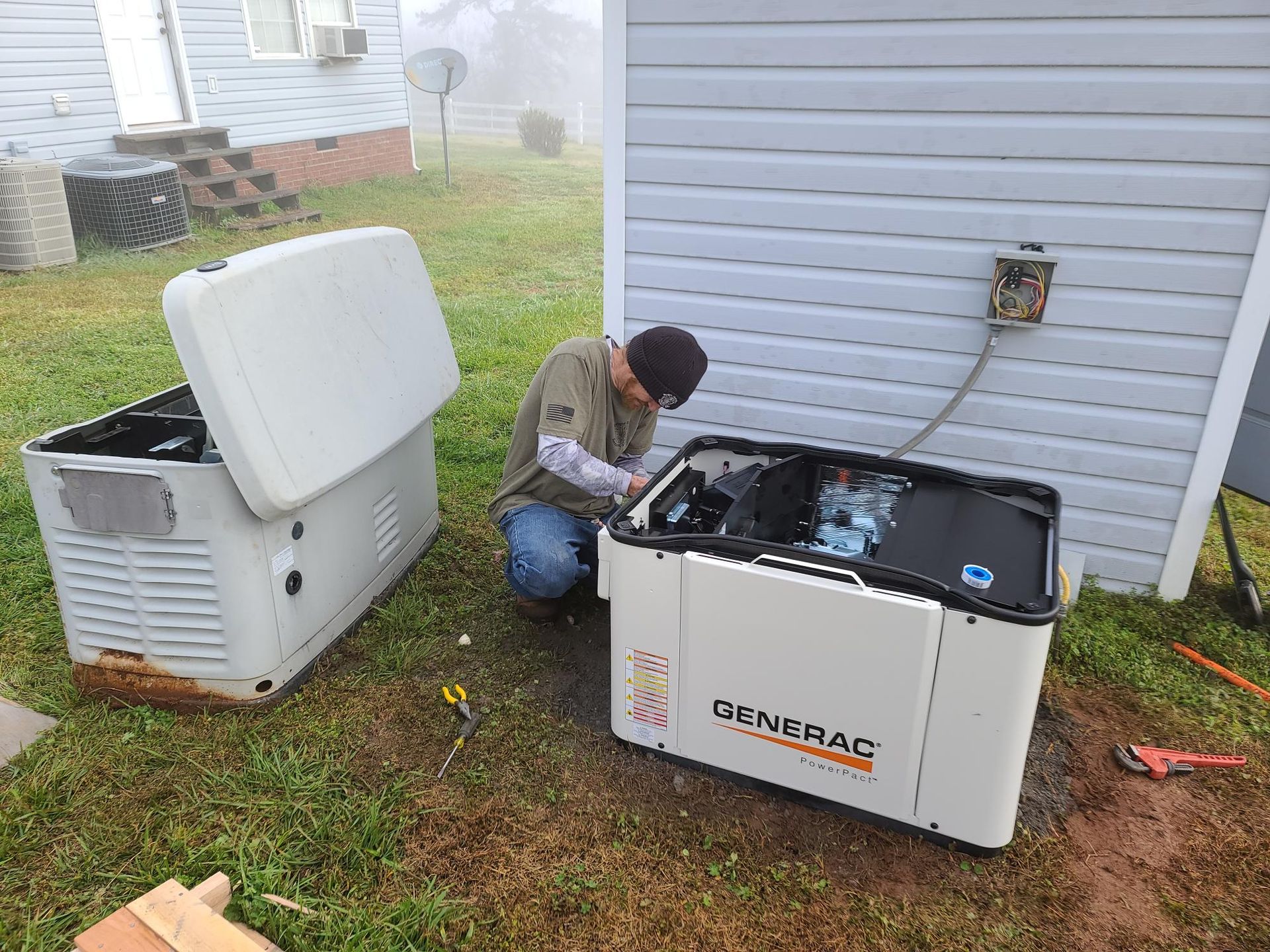 Person kneeling beside an open Generac generator, working on it outside by a house after rain.