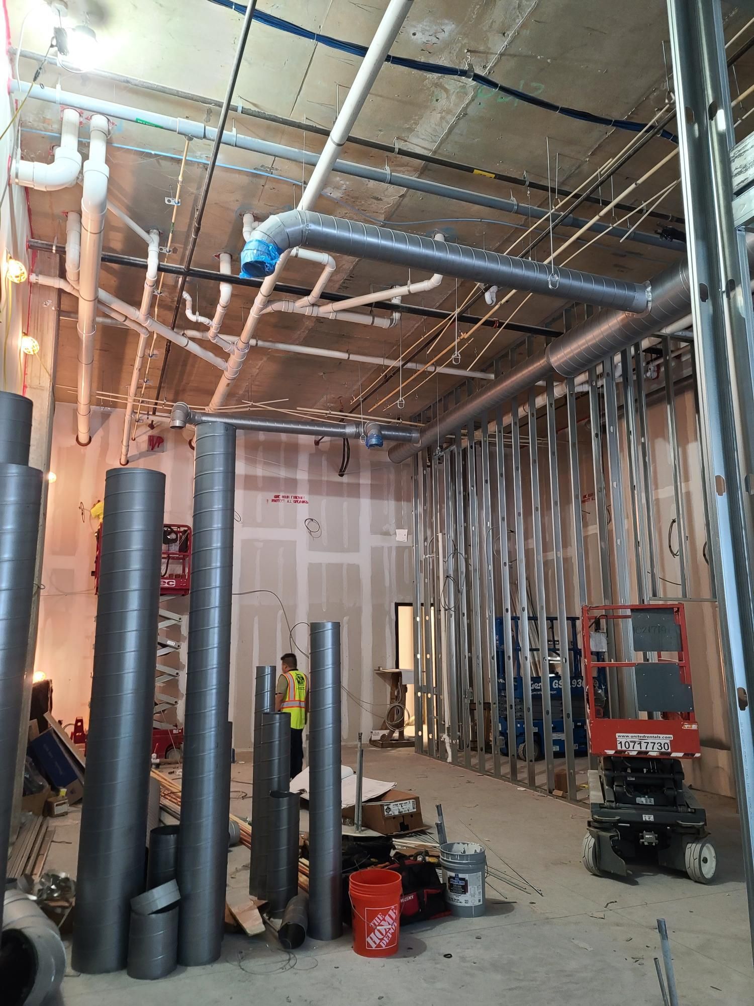 Unfinished interior with exposed pipes, insulation, tools, and a red lift in a construction workspace.
