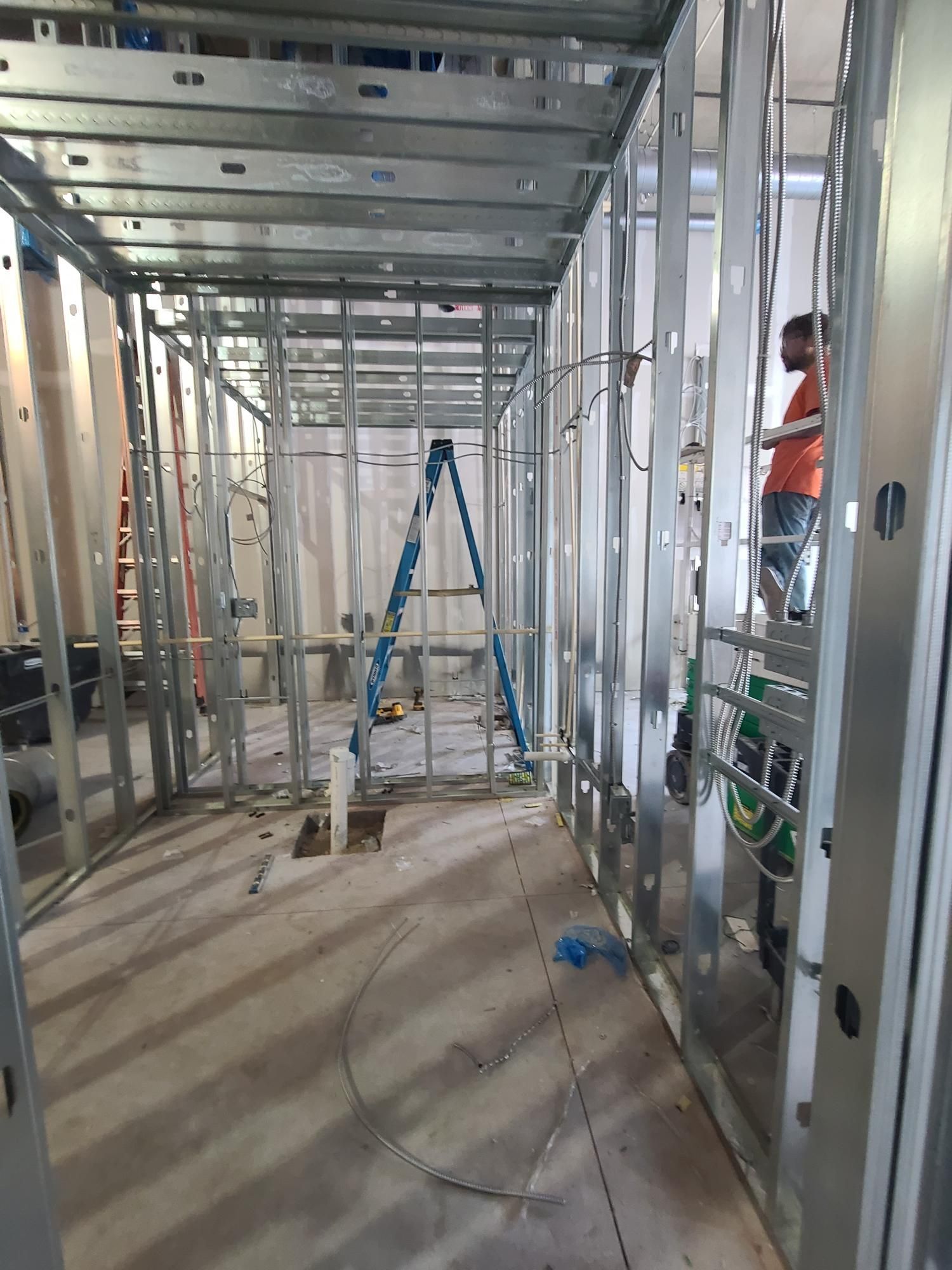 Interior of an unfinished building frame with metal studs, ladders, tools, and exposed wiring on the floor.