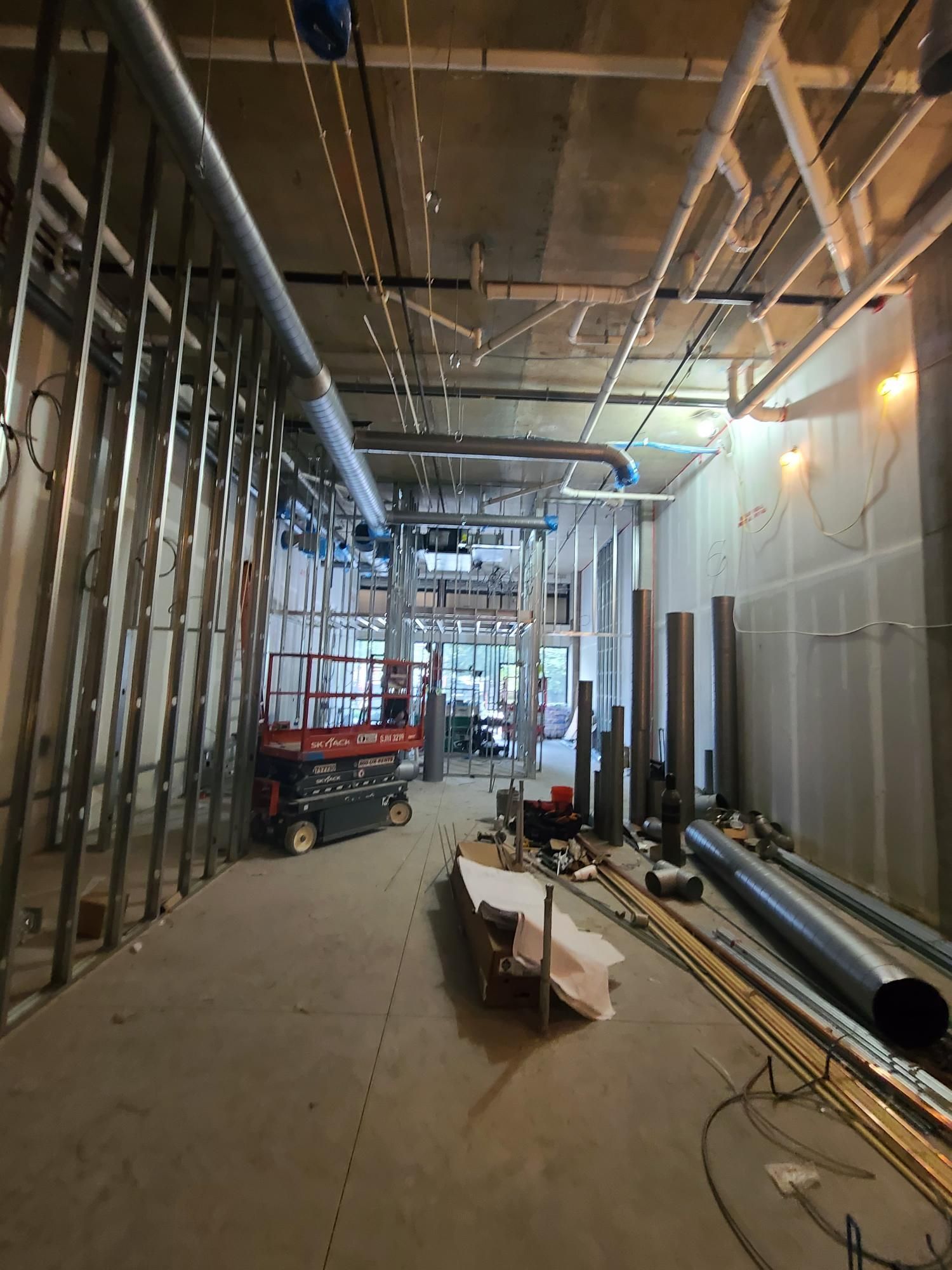 Unfinished interior construction corridor with exposed ceiling pipes, drywall, tools, and a red scissor lift.