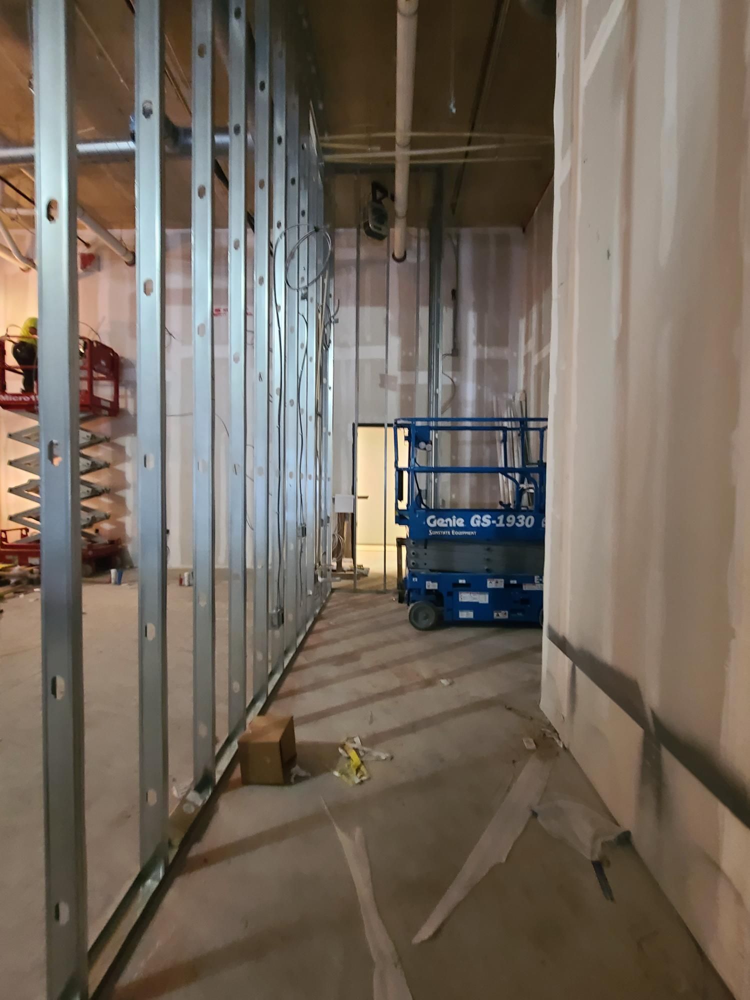 Unfinished hallway with metal studs, exposed walls, and a blue scissor lift at the far end