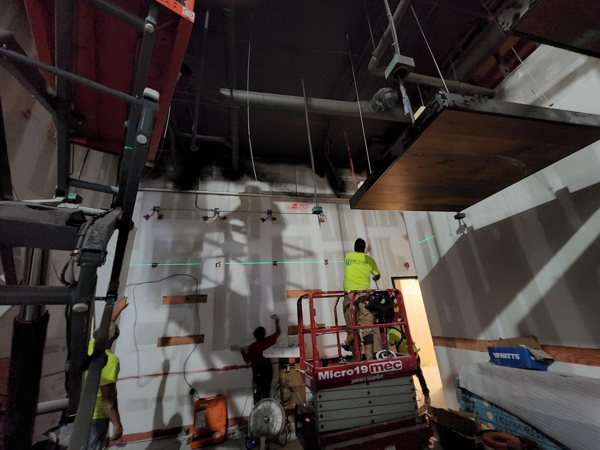Workers on lifts painting white wall panels in an industrial interior under construction.