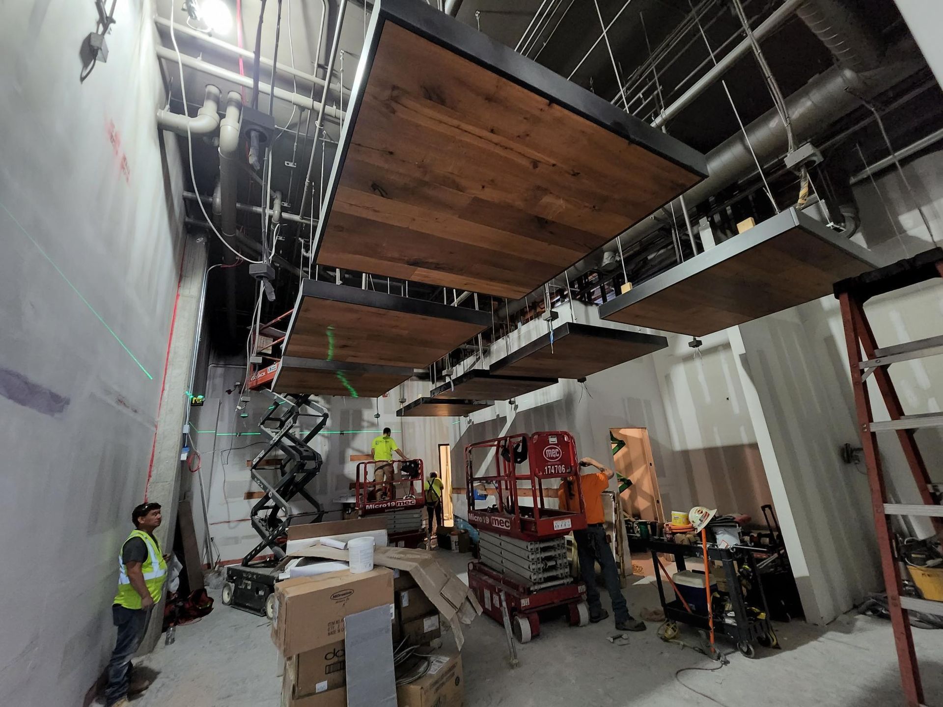 Workers on lifts install ceiling panels in a construction site with scaffolding and materials.