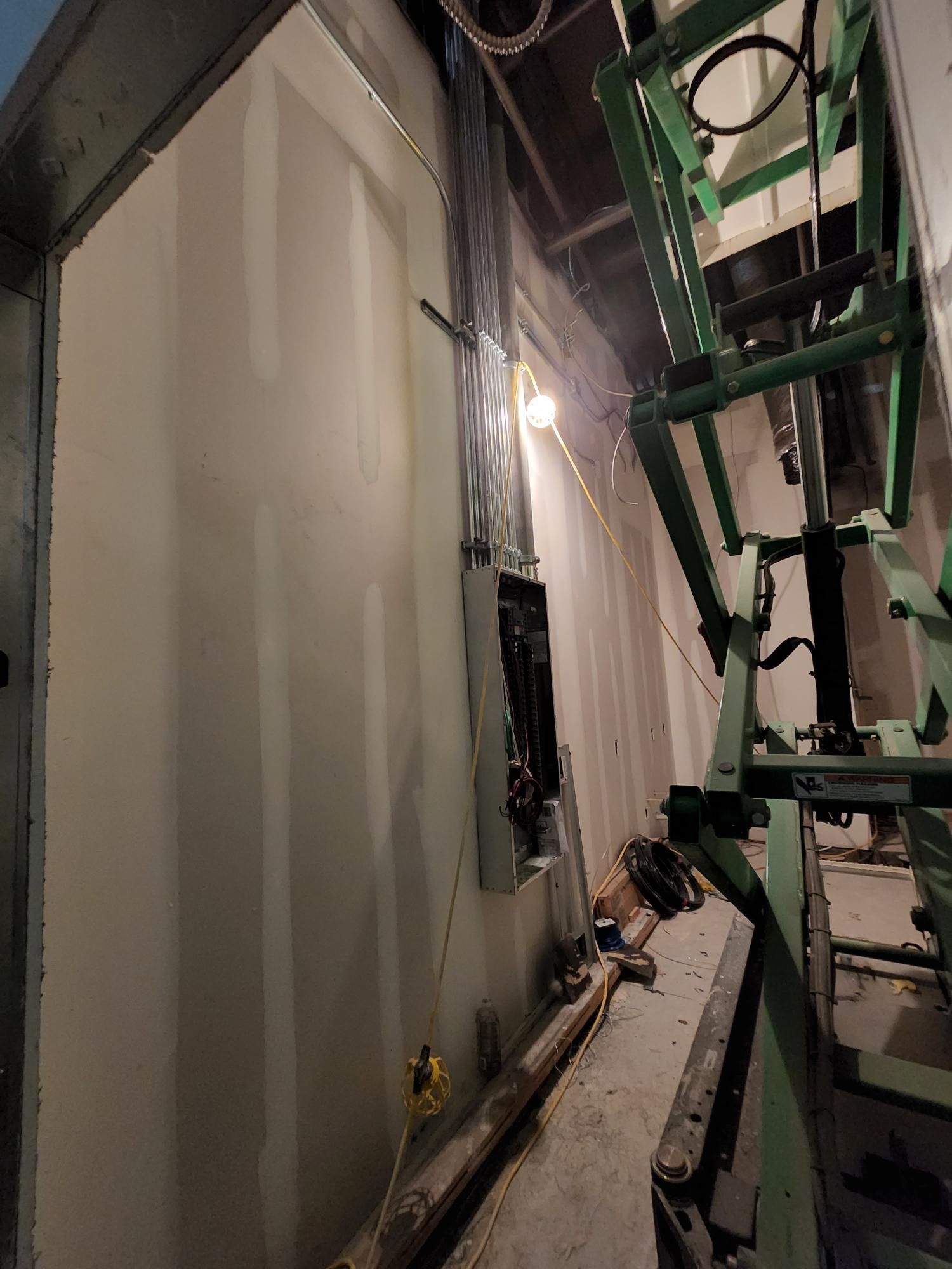 Narrow industrial corridor with white walls, exposed pipes, and green scaffolding overhead.