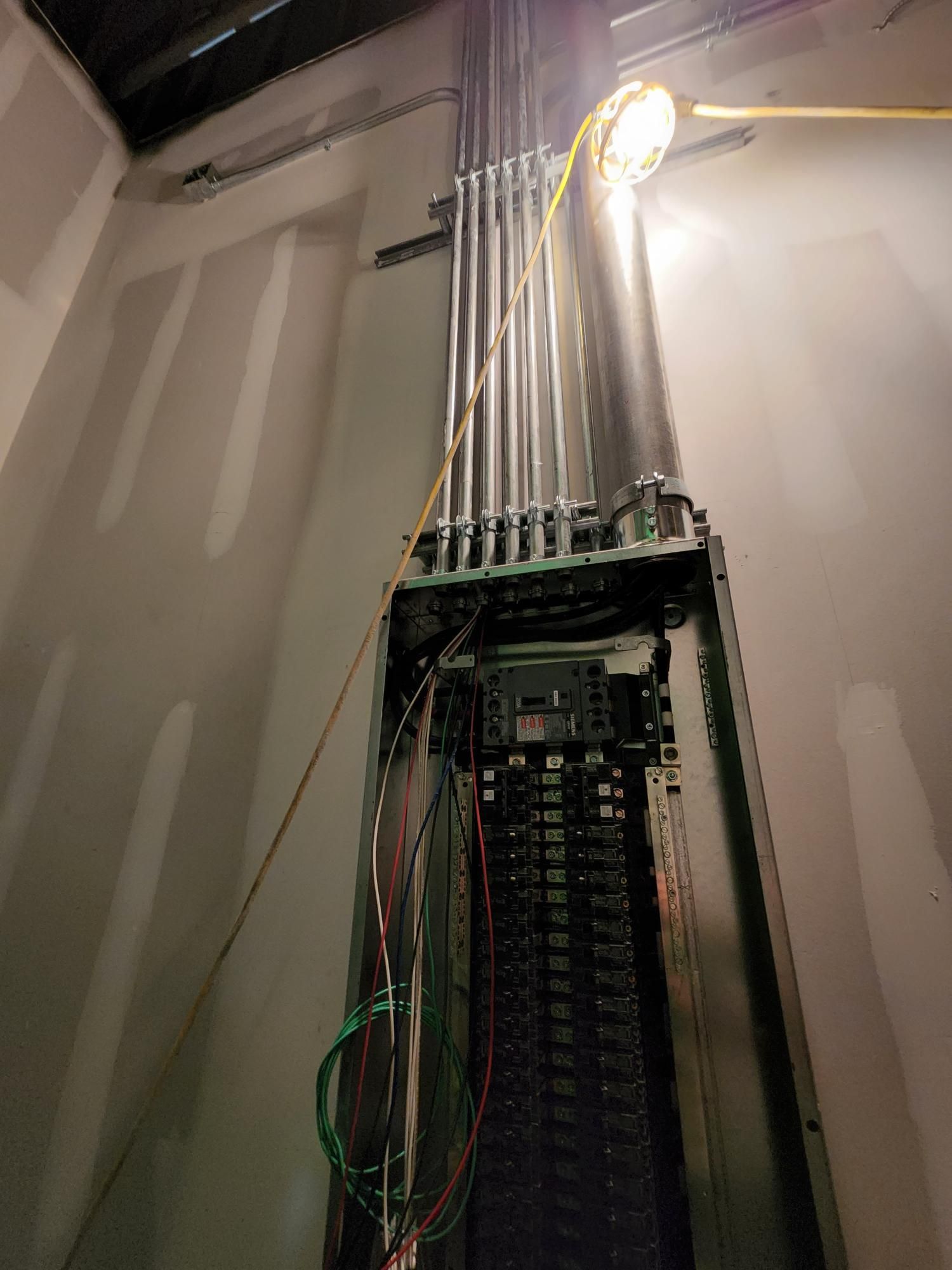Electrical panel with exposed wiring and breakers in a utility room, lit by a bright overhead bulb