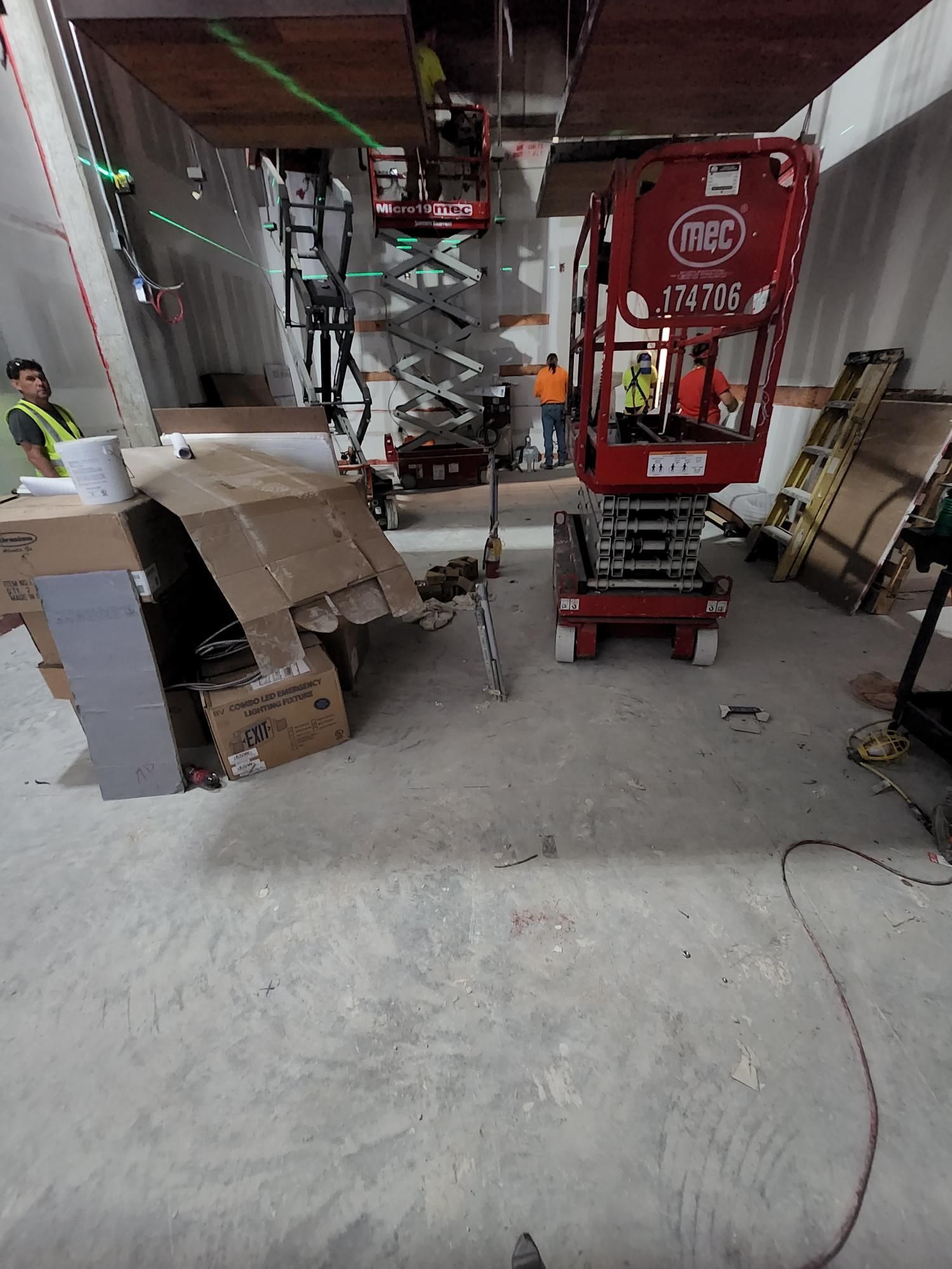 Indoor construction area with a red lift, cardboard boxes, tools, and hanging cables on a concrete floor