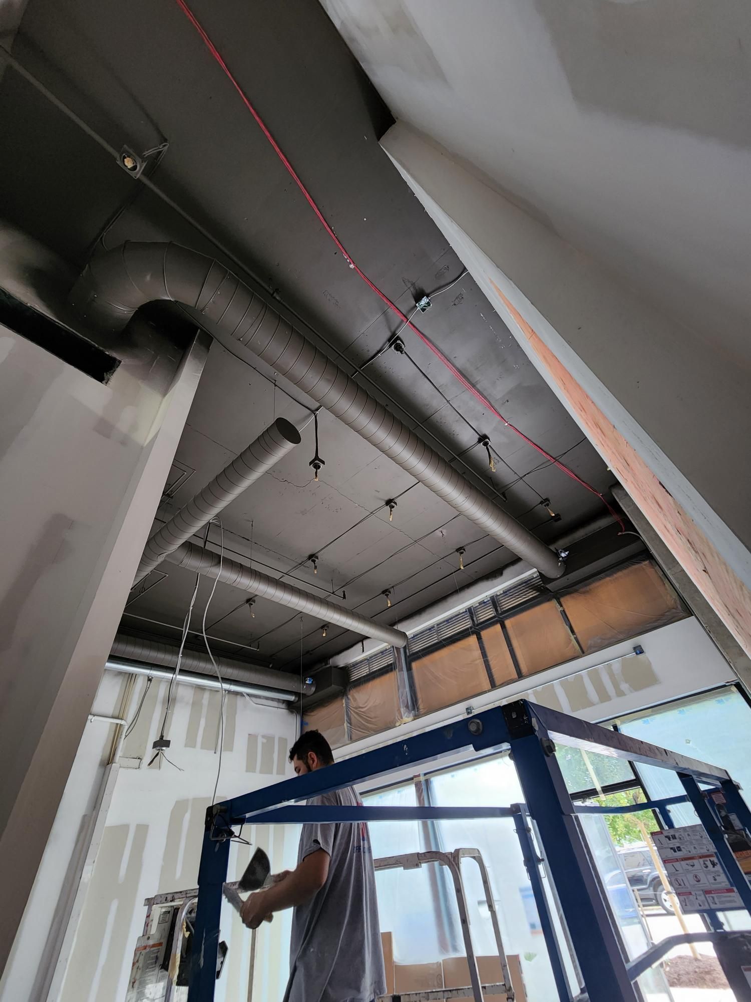 Worker installing ceiling fixtures in a partially finished room with exposed ducts and pipes.
